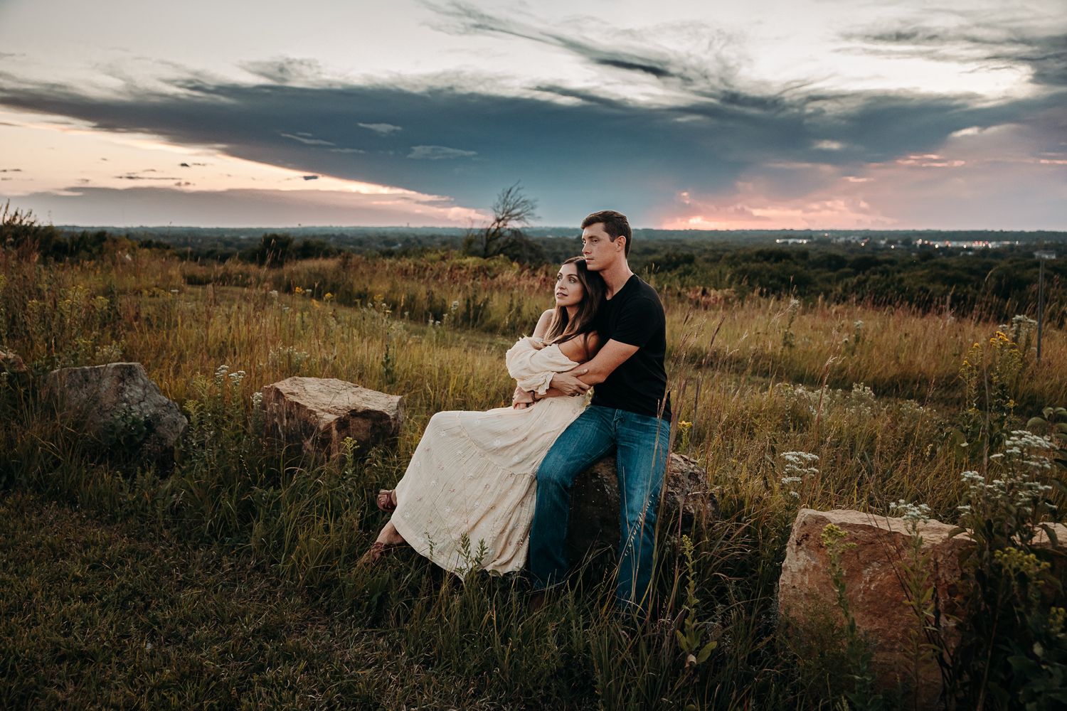 A couple shares a romantic moment sitting on a rock surrounded by wildflowers at sunset.