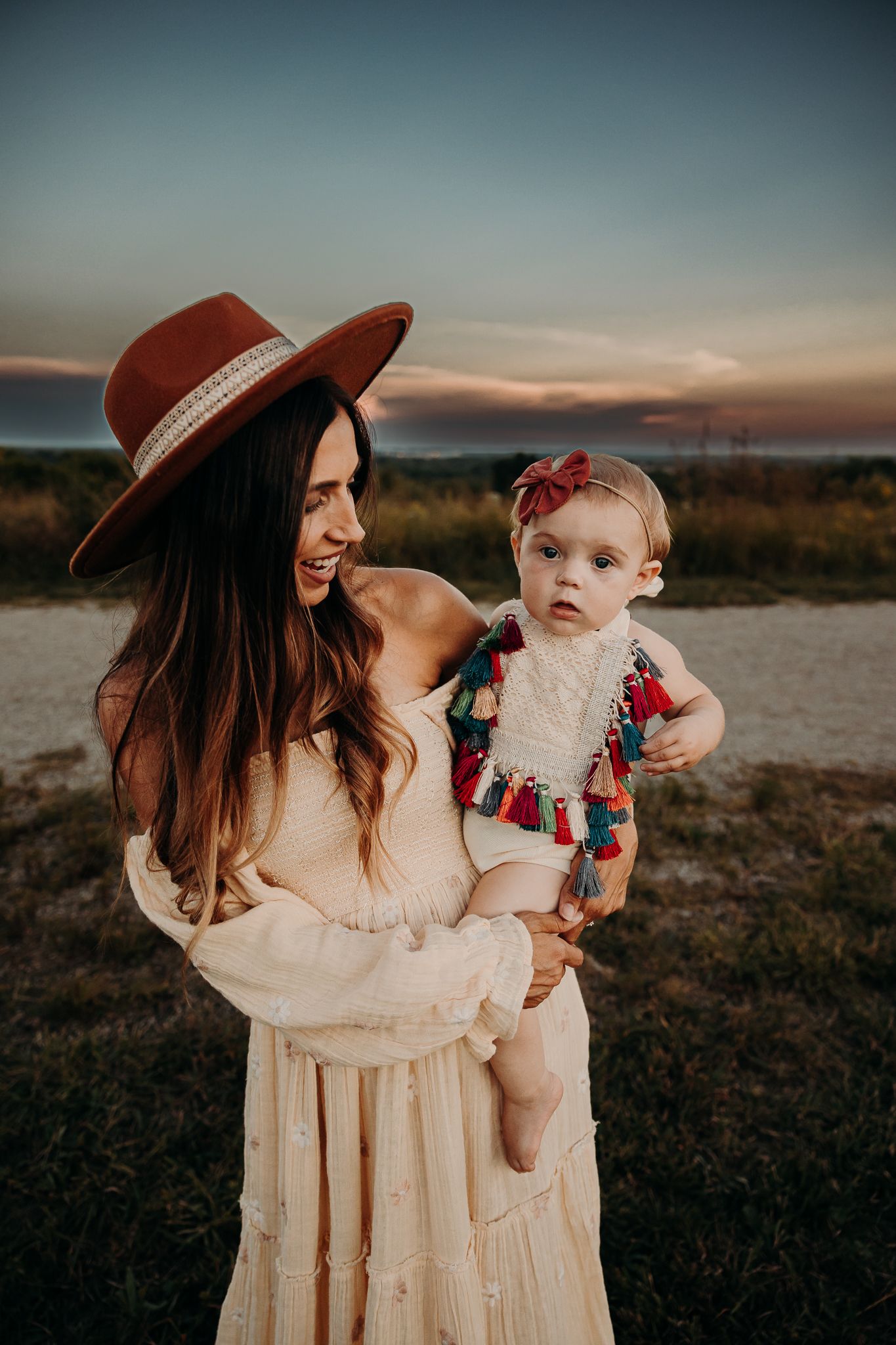 A smiling mother in a boho hat and cream dress holds her baby girl in a colorful tassel romper.