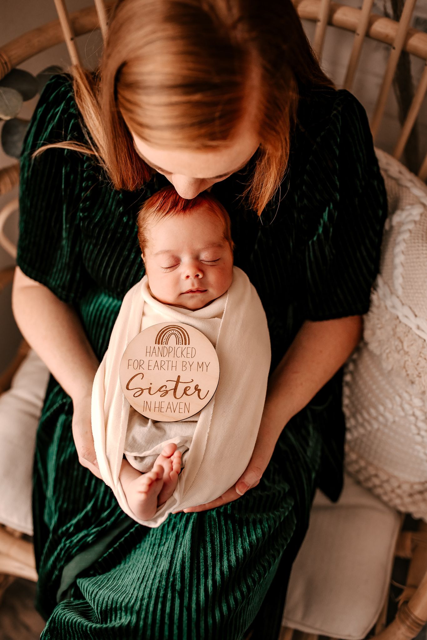 Mother in green velvet dress holds swaddled newborn with handpicked for earth by sister in heaven wooden sign.