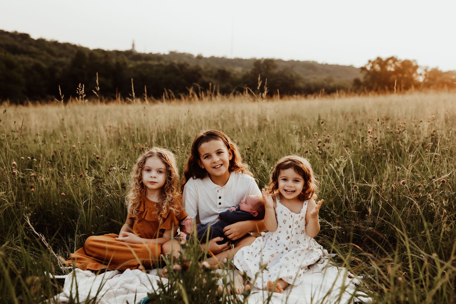 Three siblings sitting in golden meadow field together with oldest holding newborn baby sibling.