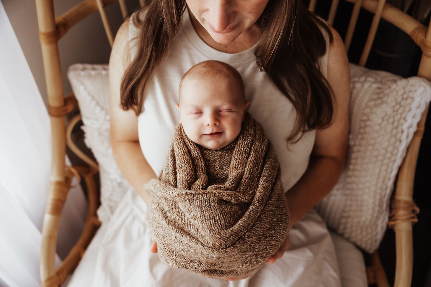 Mother holding swaddled smiling newborn baby in brown knit wrap while sitting in a rattan chair.