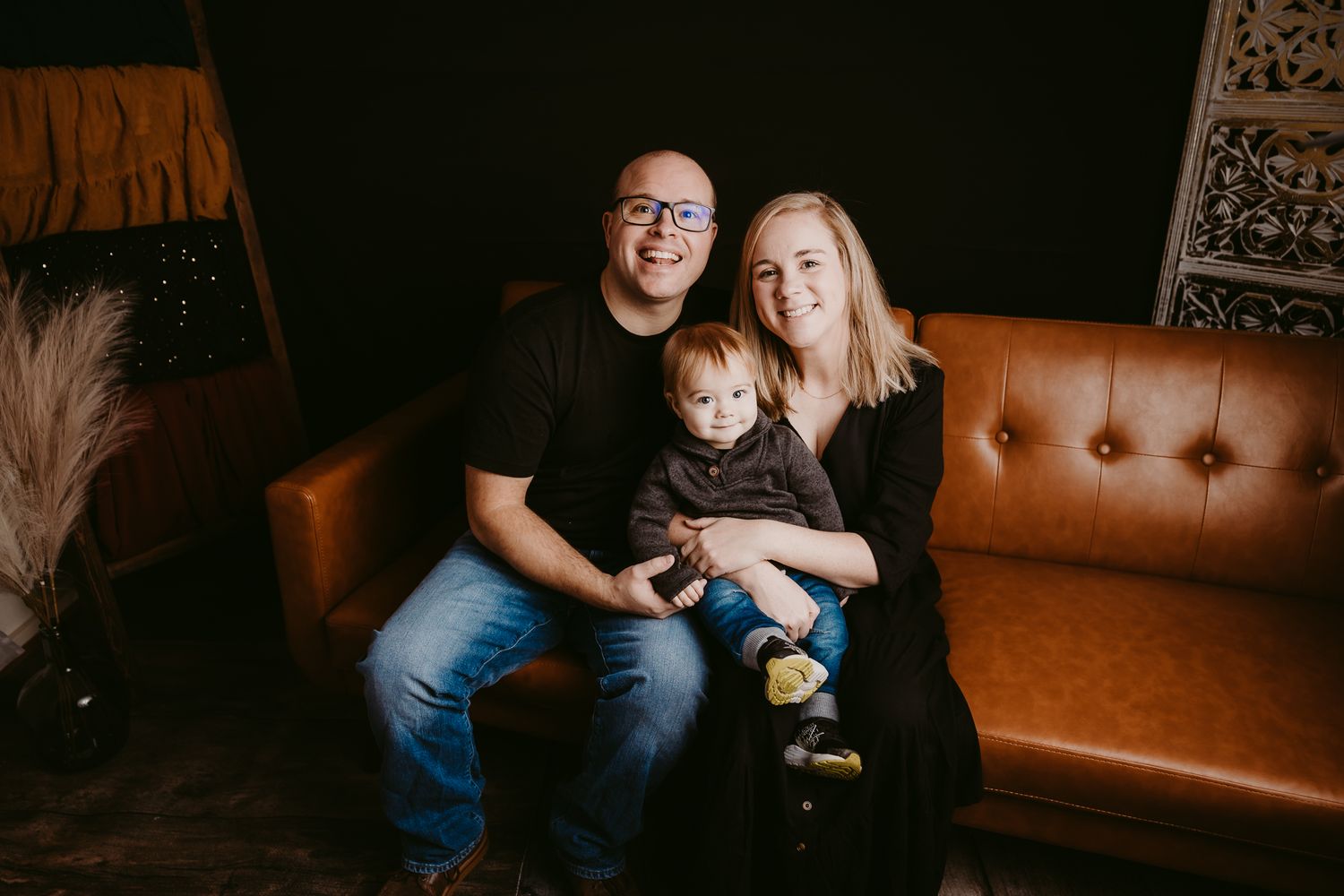 Happy family of three poses on a brown leather couch with their toddler between them.