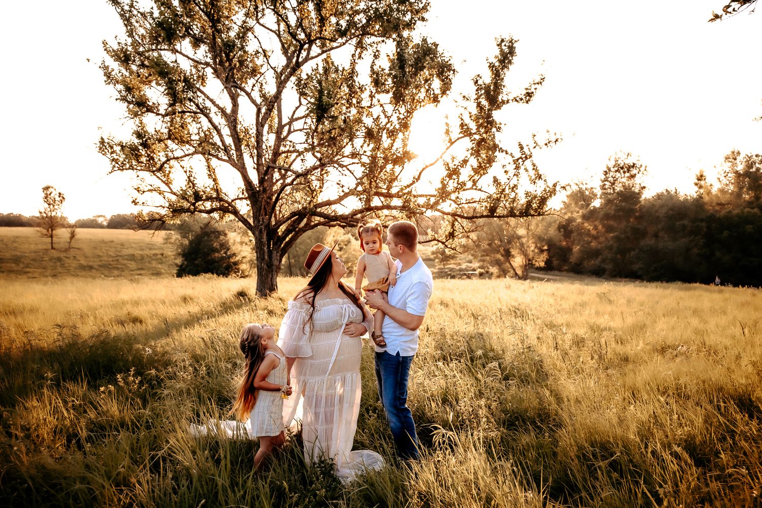 A family of four enjoys a golden hour moment together in a sunlit meadow beneath a large tree.