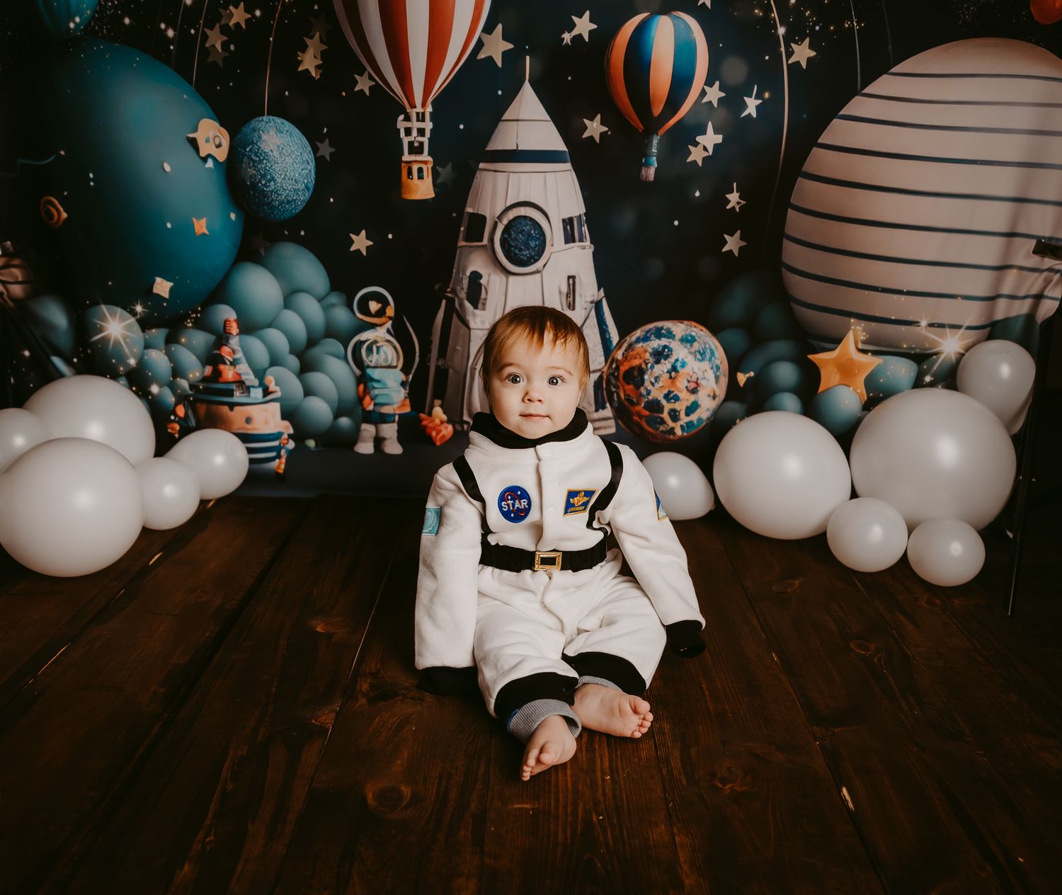 Cute toddler in NASA astronaut costume sits among space-themed balloons and rocket ship backdrop.
