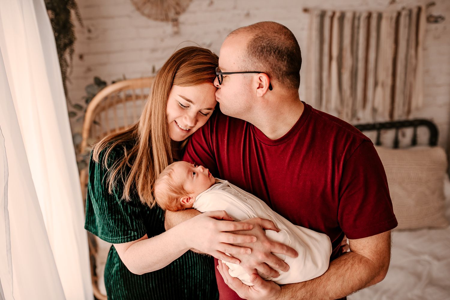 New parents share a tender moment kissing over their swaddled newborn baby in a cozy bedroom setting.