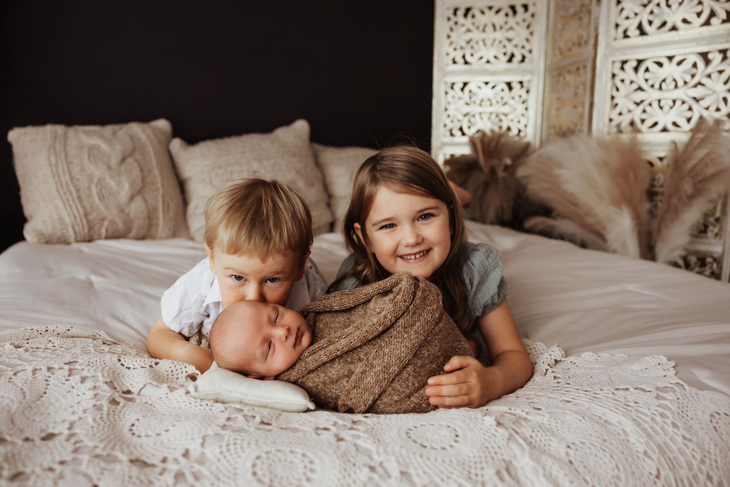 Two smiling siblings cuddling newborn baby on white bedding.