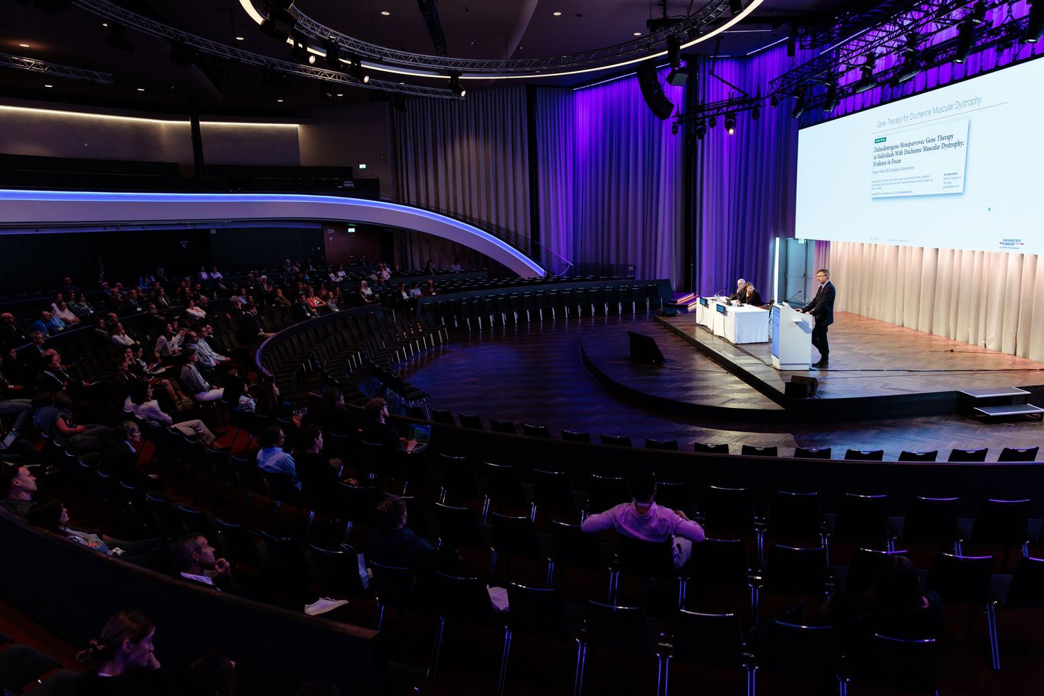 A large auditorium filled with attendees watching a presenter on stage with a illuminated screen at a professional conference.