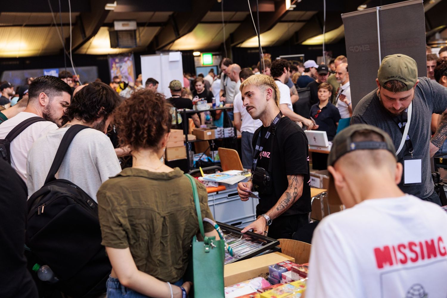 Crowded indoor market event with tattooed vendors and diverse attendees browsing tables filled with colorful merchandise.