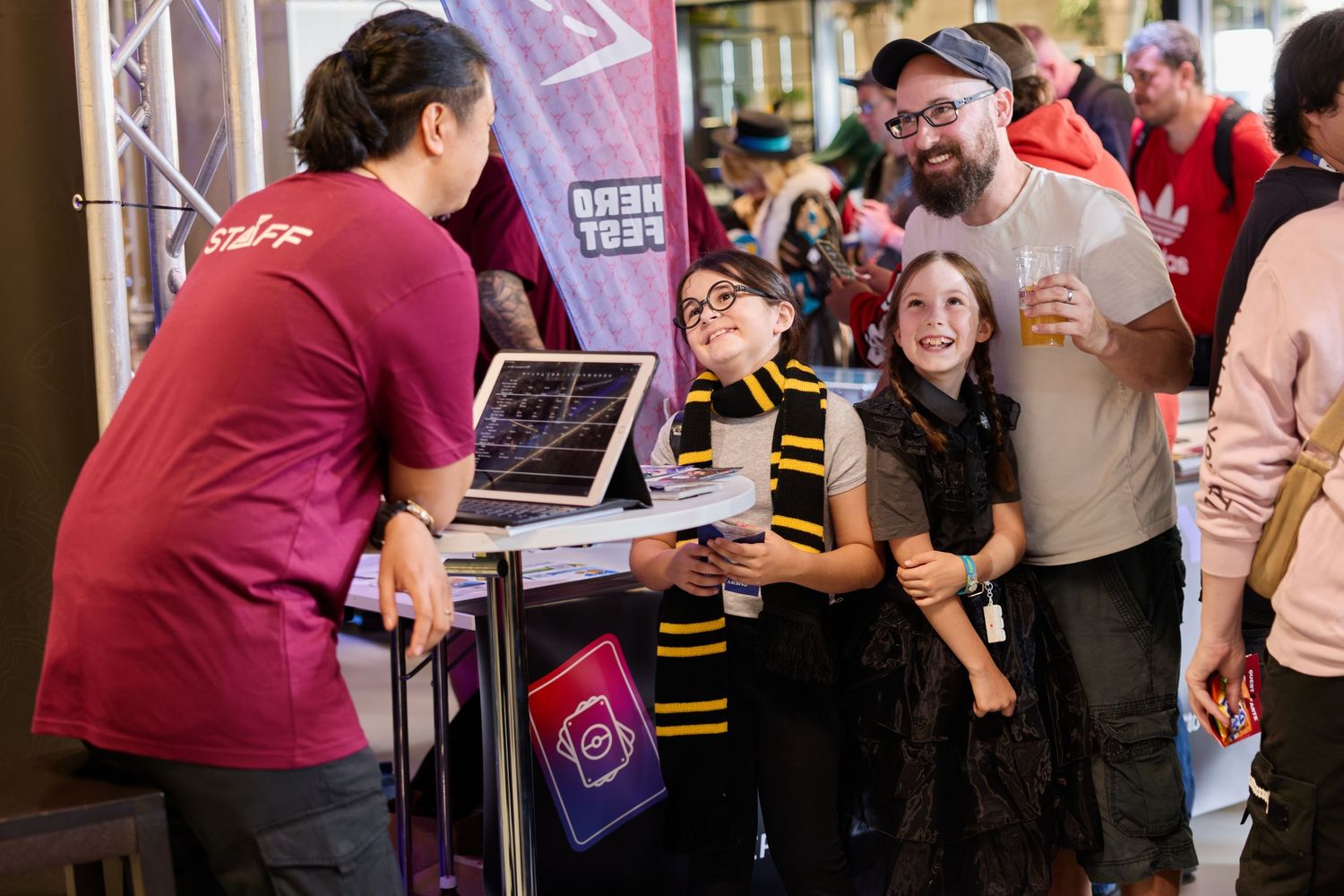 Group of visitors smiling at a staff member behind the Erupt TCG booth at HeroFest with a laptop and display cards visible on the table