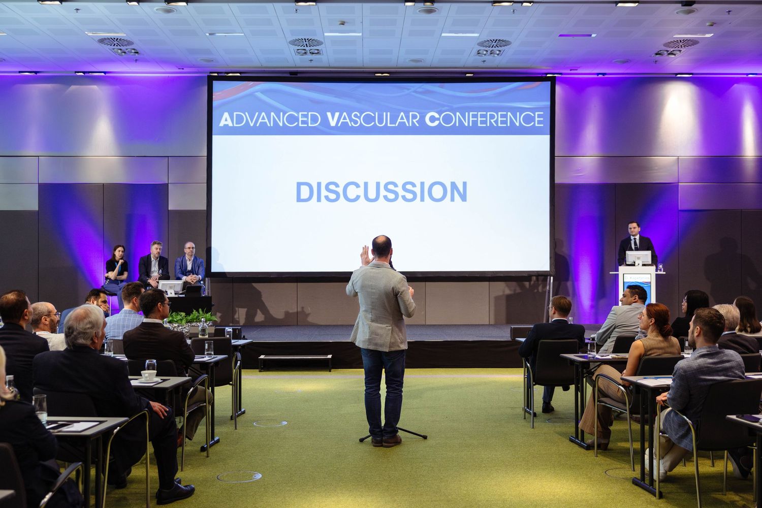 Attendees at the Advanced Vascular Conference engage in a discussion session in a modern conference hall with a large projection screen.