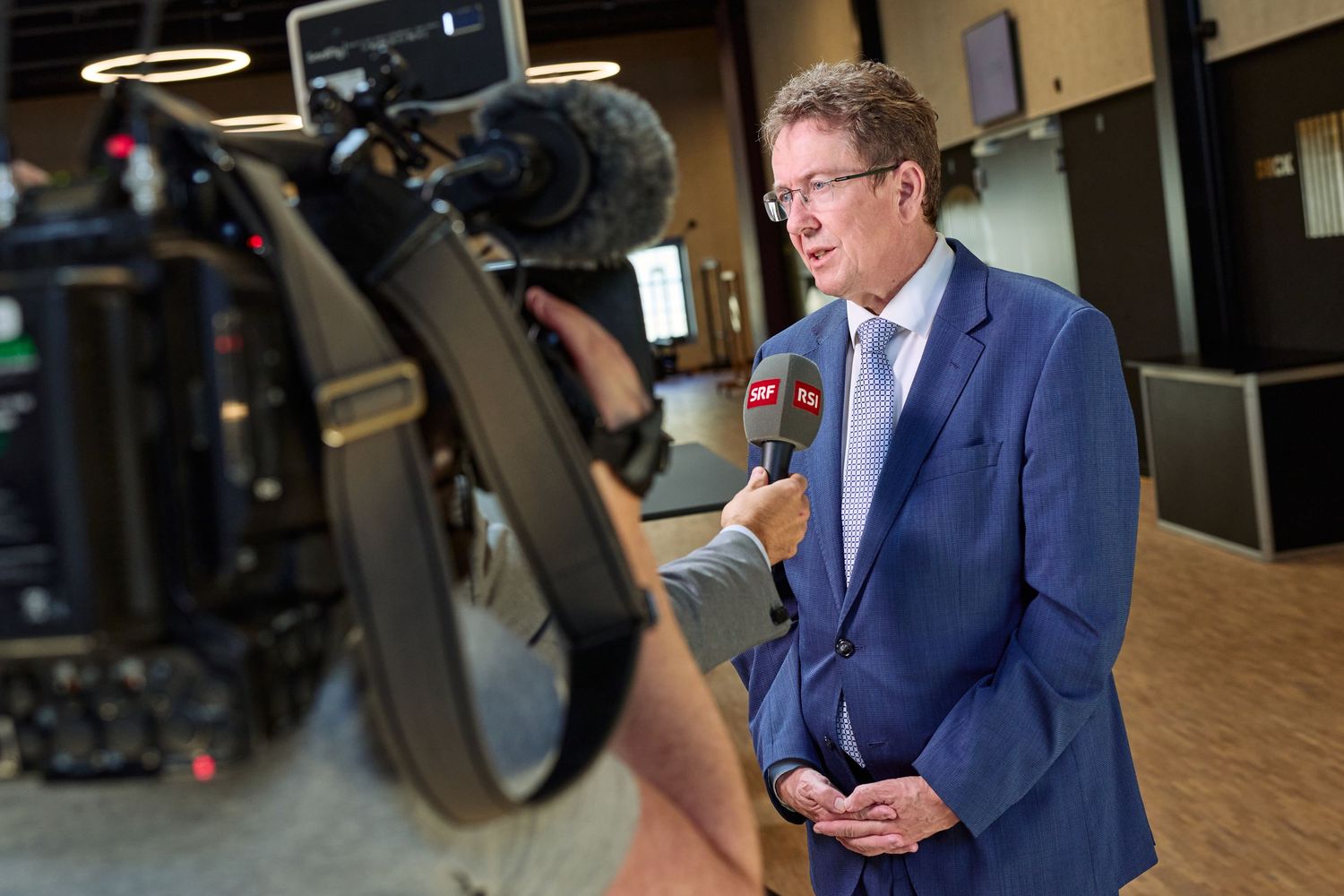 Albert Rösti is interviewed by an SRF journalist, speaking into a handheld microphone during an event.