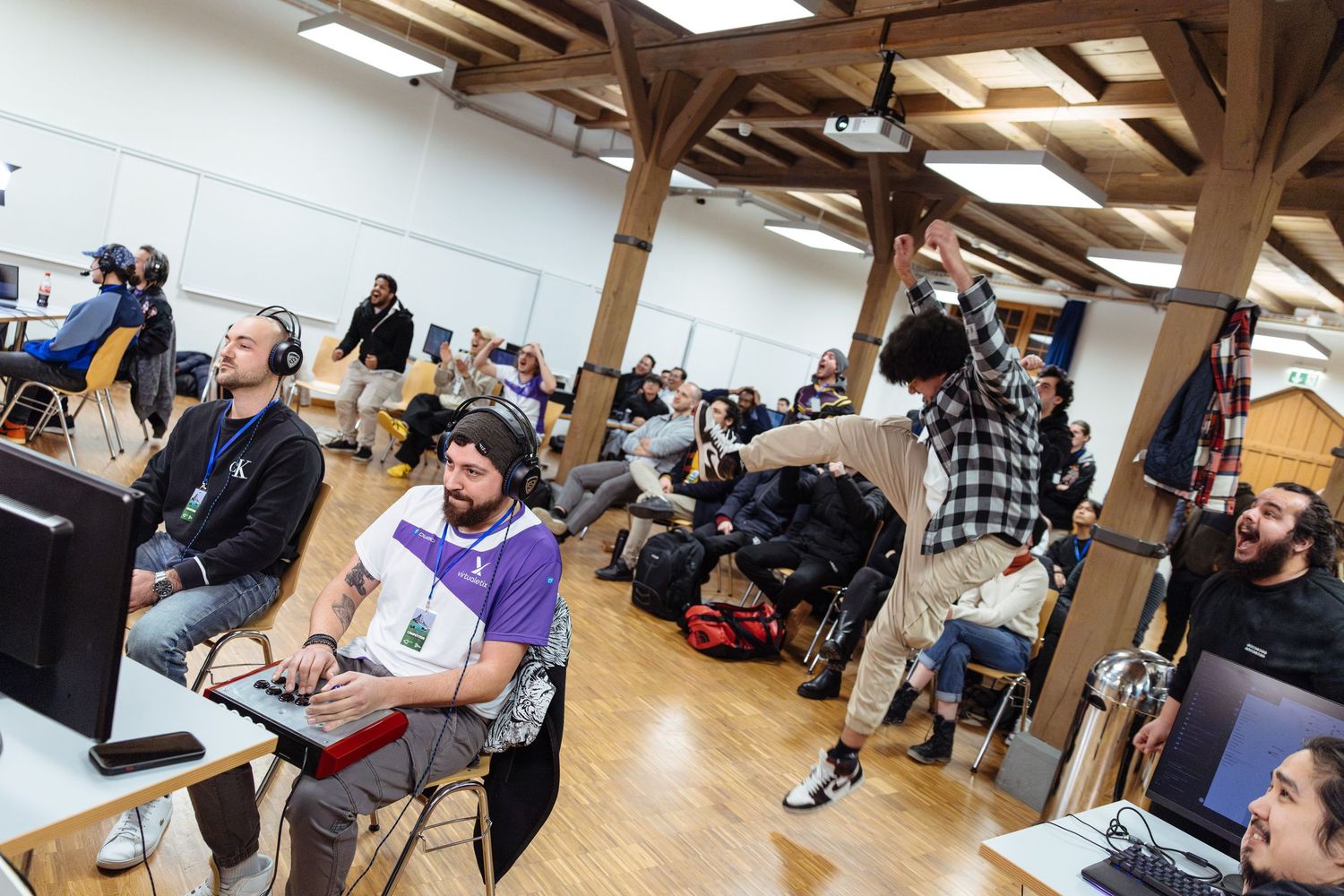 Ragebrownie jumps and cheers after Chucky-Z wins his match at Hidden Retreat, a Tekken tournament in a wood-beamed hall.