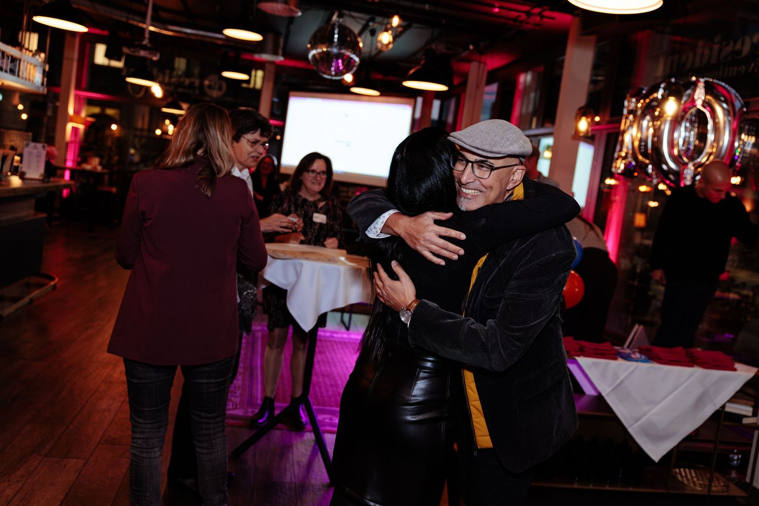 Two people share a warm hug at a lively bar event with pink lighting and guests mingling in the background.