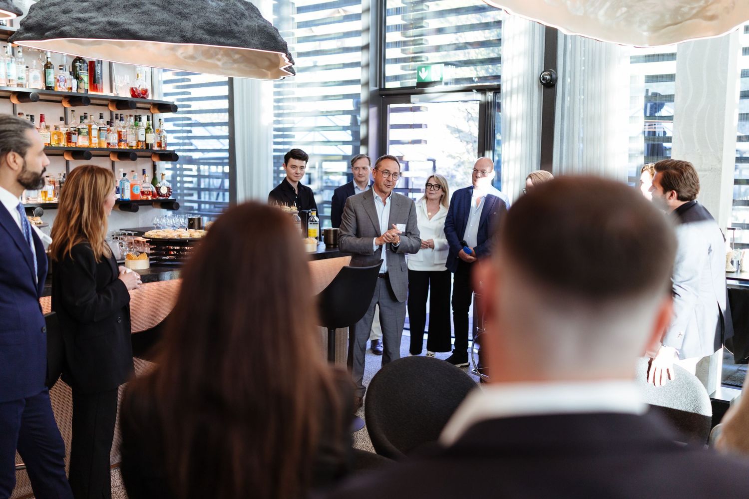 Bernard Lukey of Maison Solignac addresses business professionals at Hotel des Horlogers in Le Brassus, with attendees gathered at the bar.