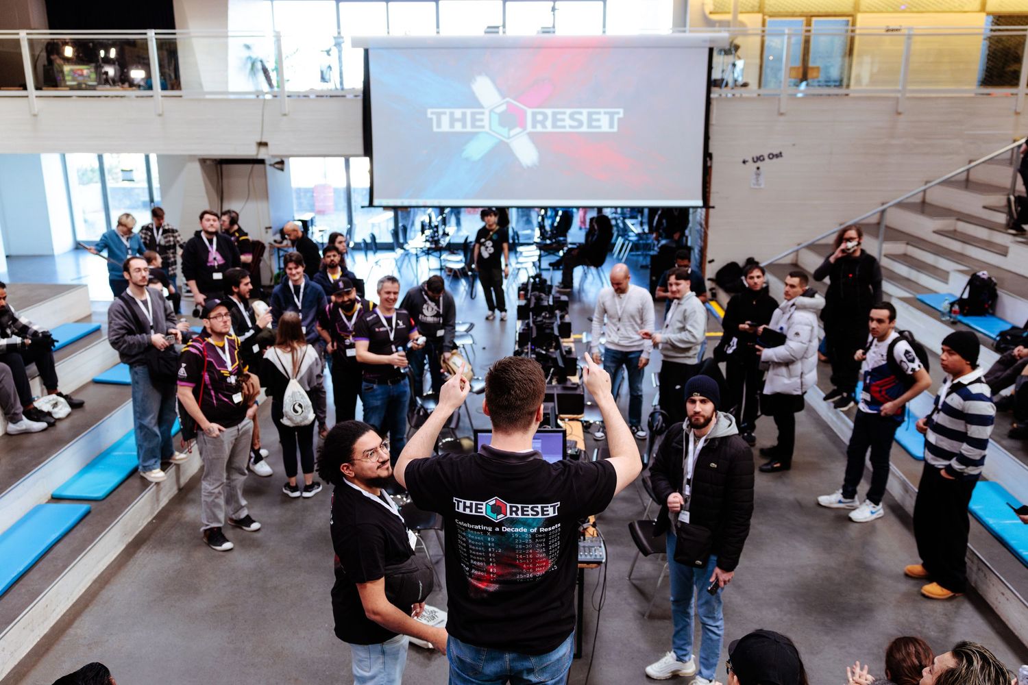 Beni tournament admin stands with raised arms addressing a group of attendees in front of a large 'THE RESET' screen at The Reset X event.