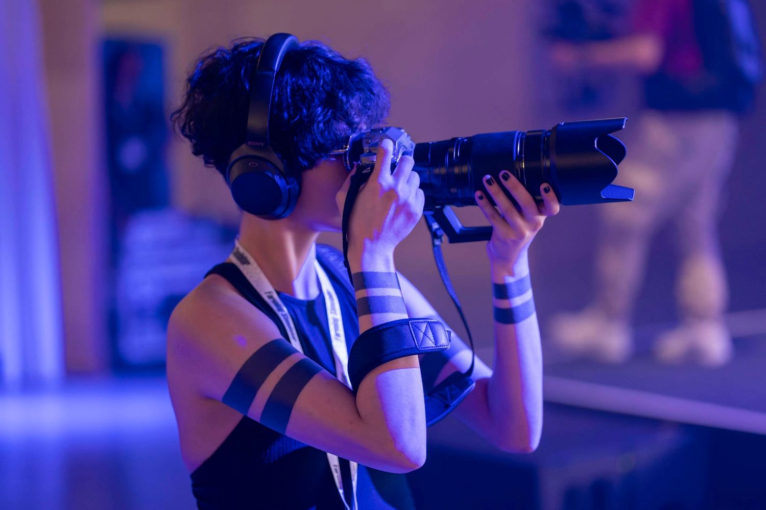 The photographer Michelle Alexis Reber also known as Lycrus with headphones capturing shots using a professional camera at a blue-lit event.