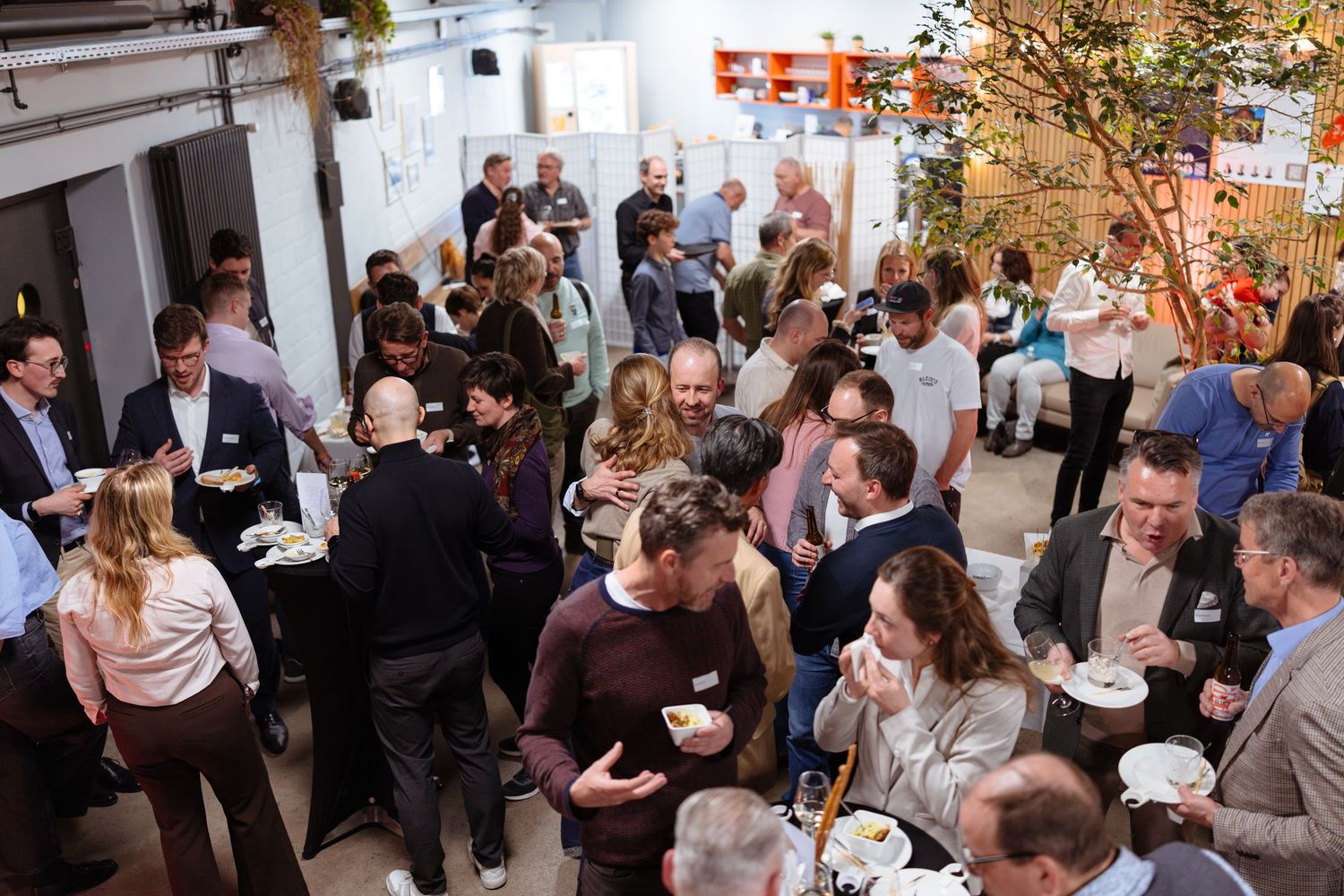 A lively indoor networking event with guests mingling, eating, and socializing in a modern venue with greenery.