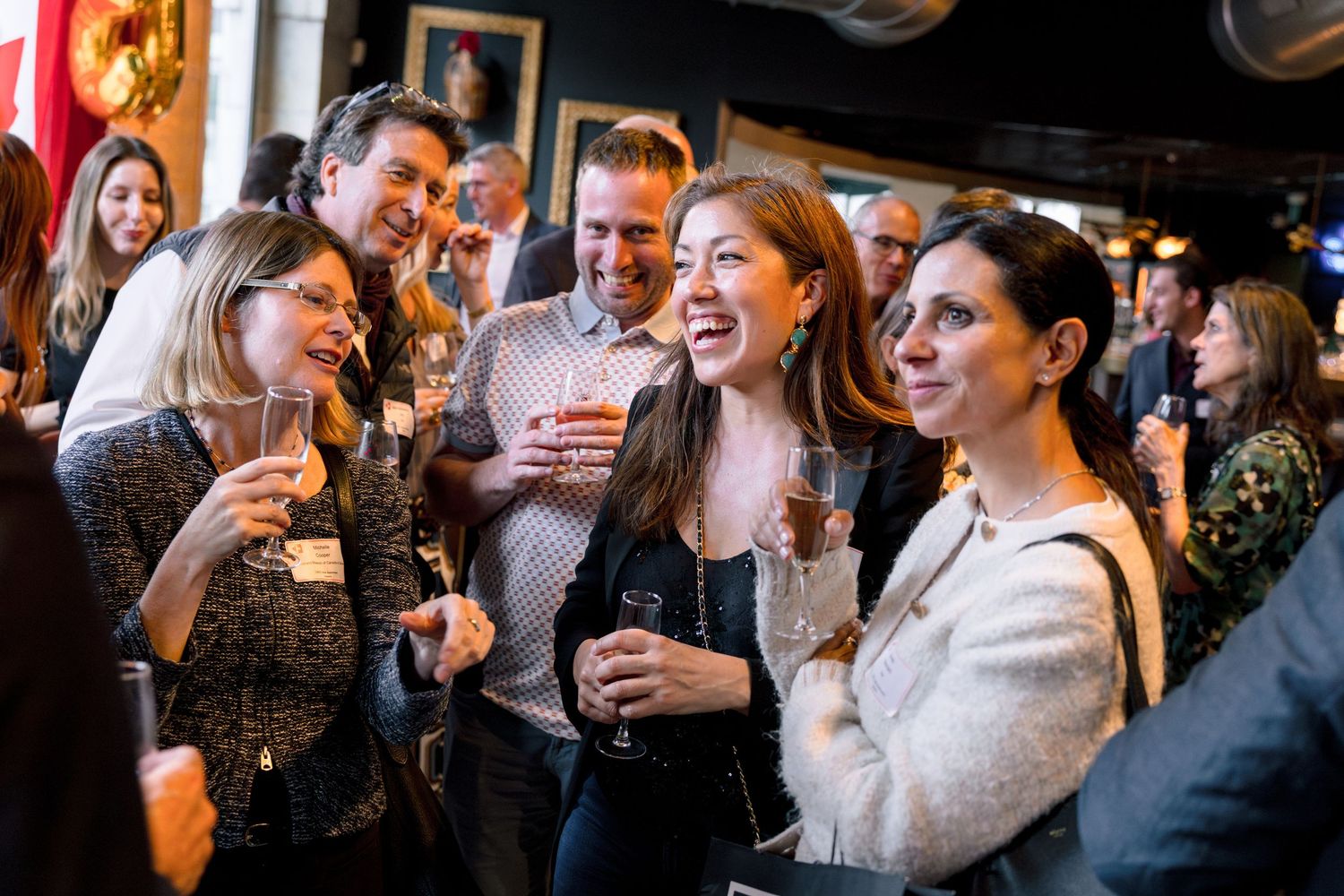 Group of professionals socializing at a networking event, laughing and holding drinks in a warmly lit venue.