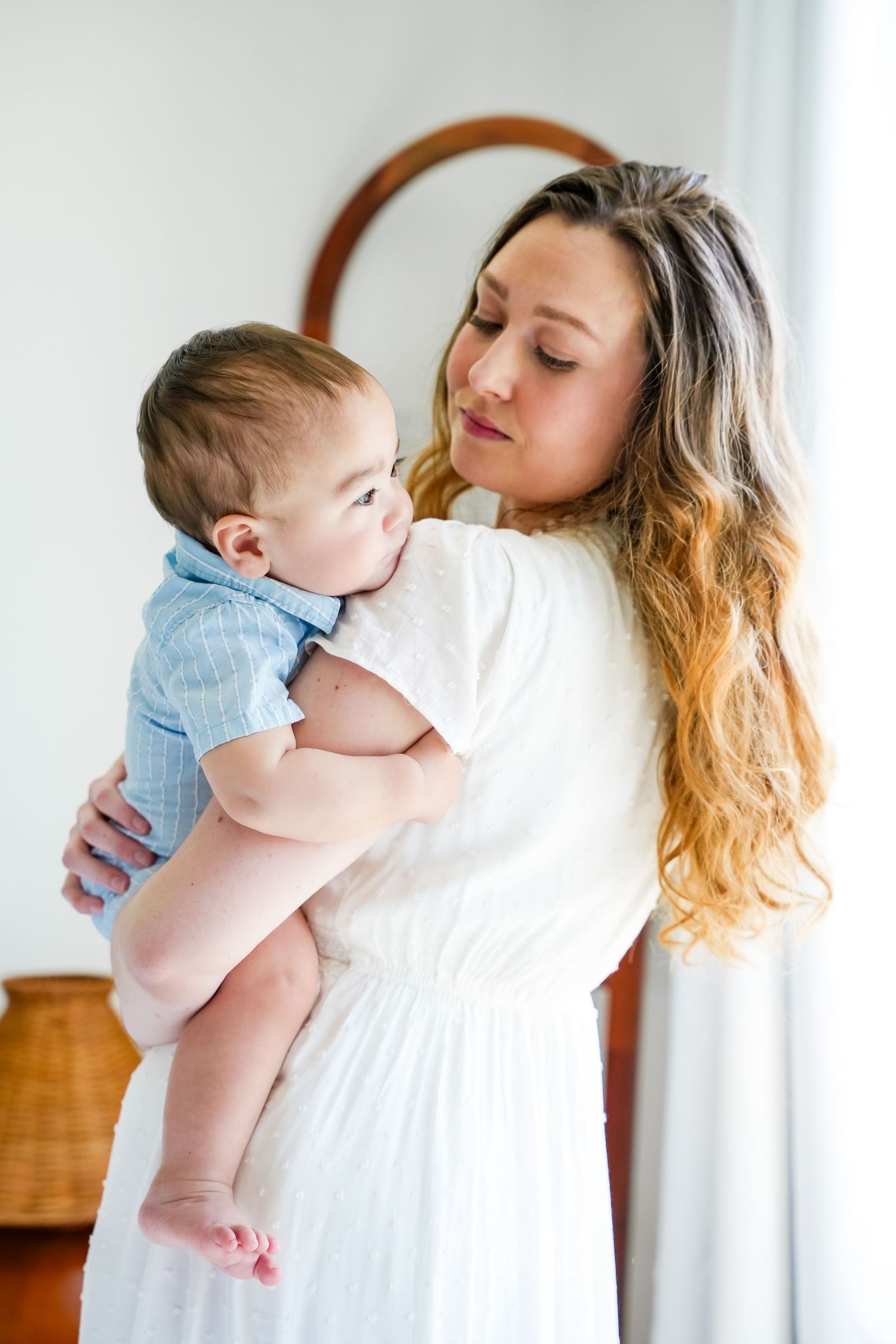 A mother in a white dress tenderly holds her baby boy in a blue outfit in a bright, airy room.