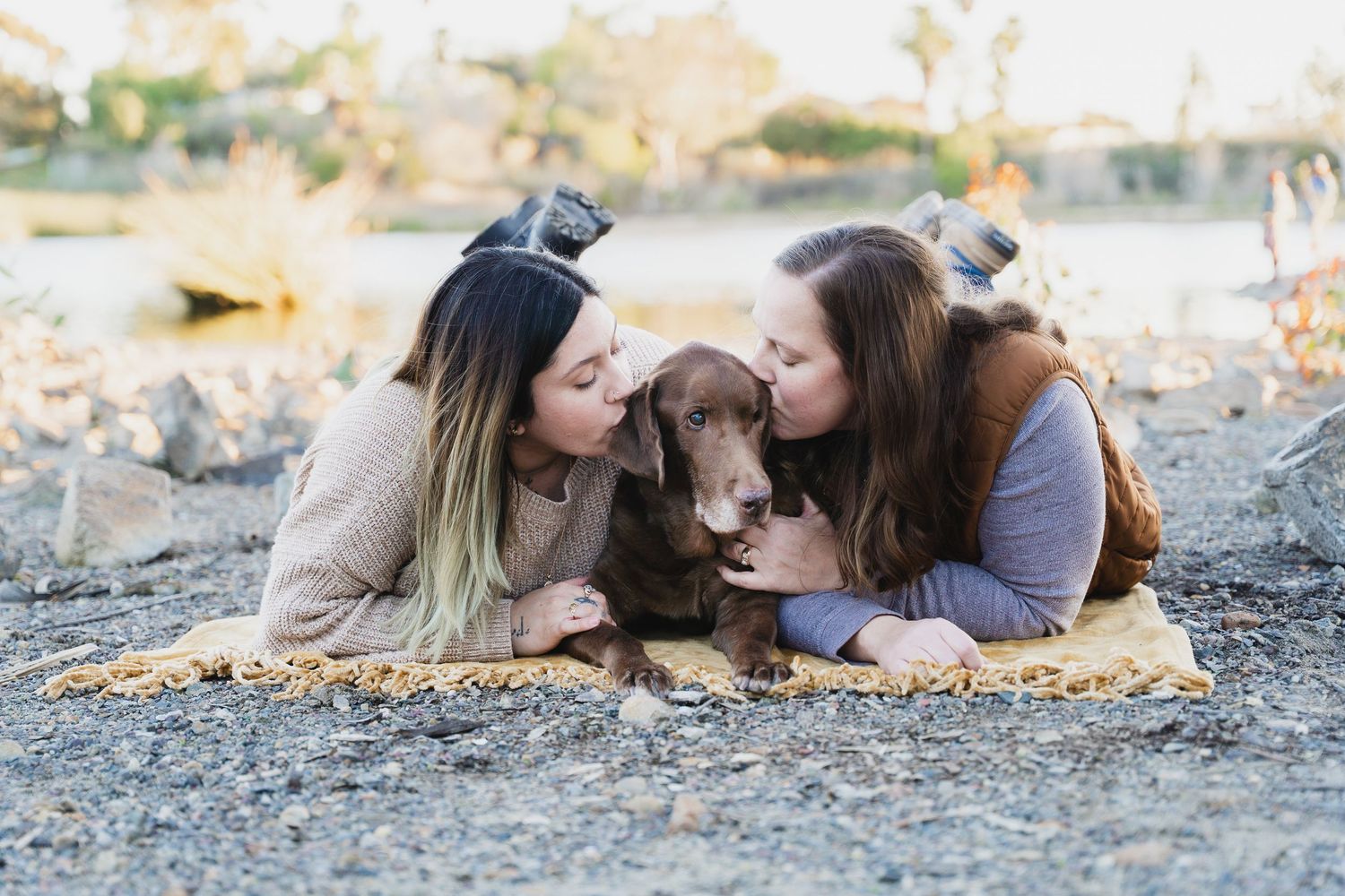 Two women kiss a chocolate dachshund on a rocky beach blanket outdoors.
