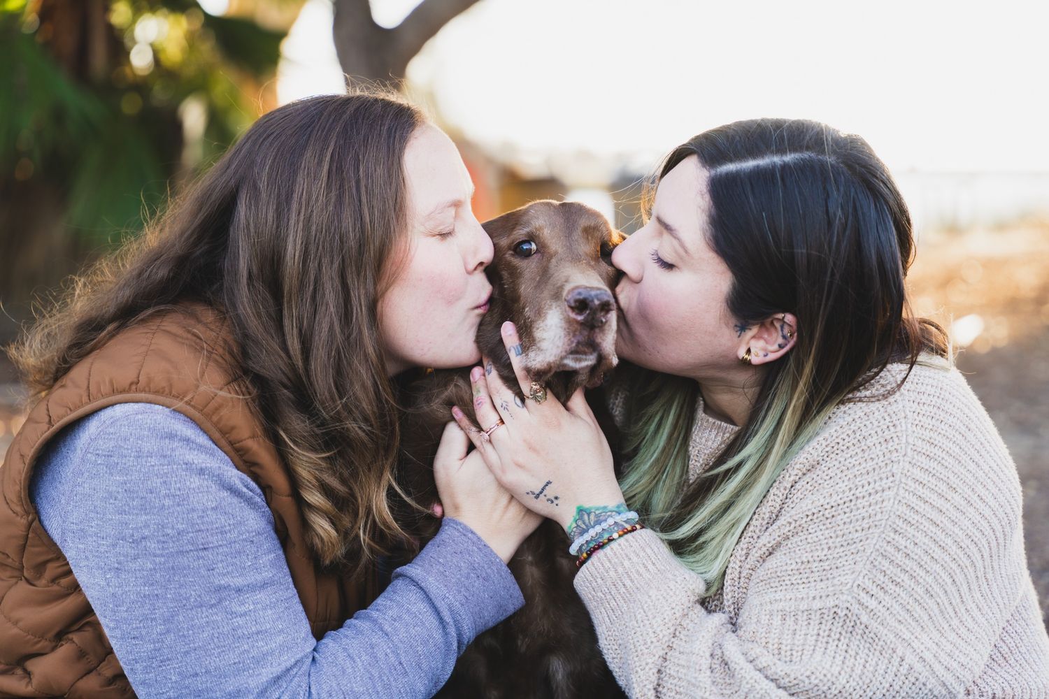 Two women kissing a brown dog on the cheeks outdoors at golden hour.