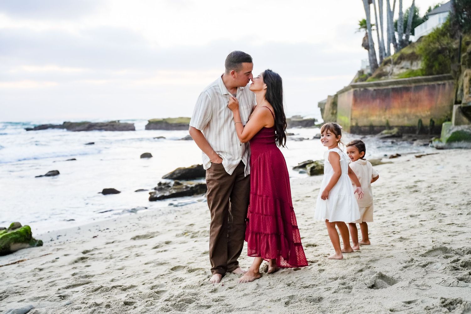 Family beach portrait with couple kissing while two young daughters stand nearby on sandy shore.