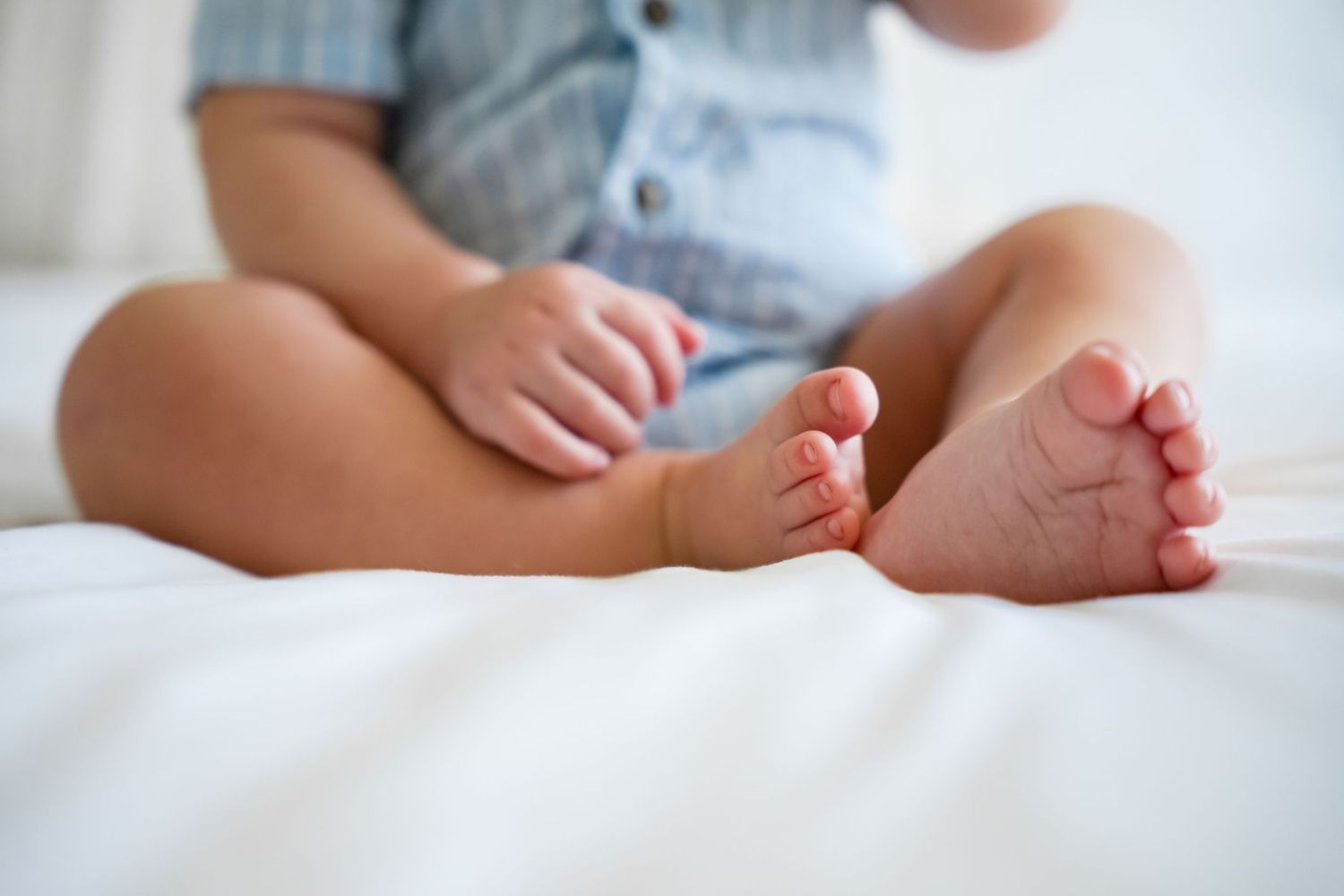 Close-up of a baby sitting cross-legged on white bedding, showing tiny feet and hands in a blue outfit.