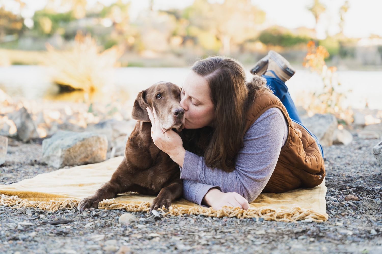 Woman lying on a yellow blanket outdoors kissing her chocolate Labrador dog near rocky terrain.