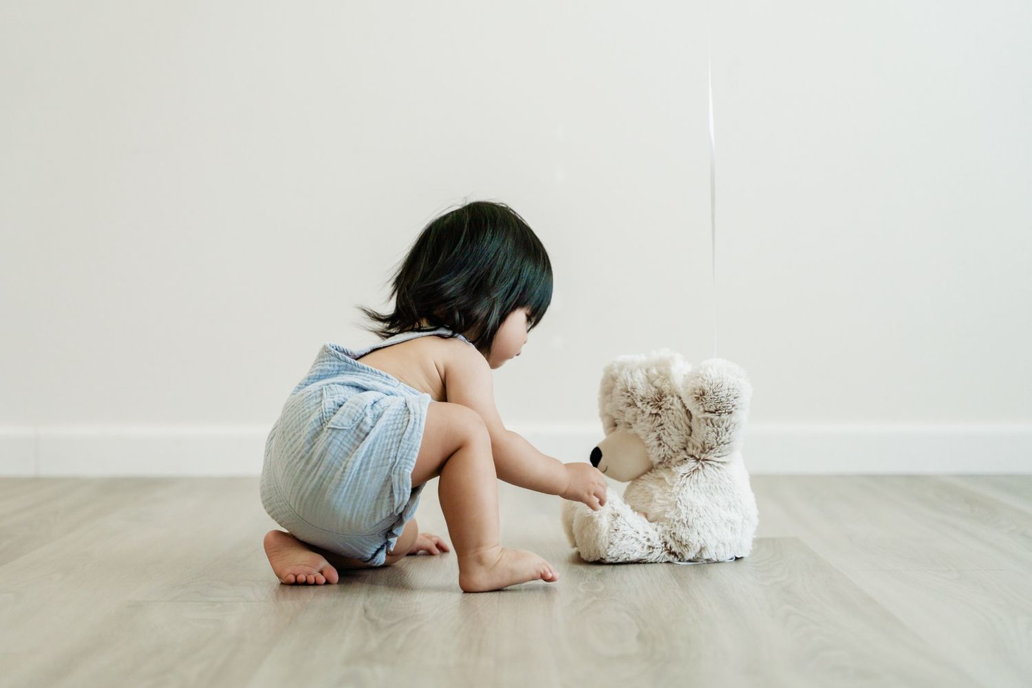 Toddler girl sitting on hardwood floor playing with a white teddy bear against a white wall.