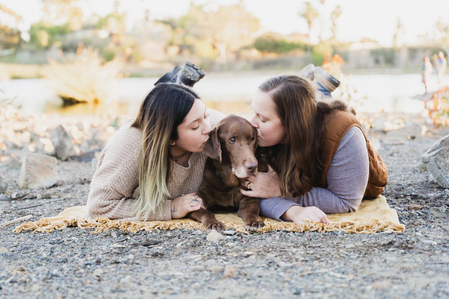 Two women lying on a blanket outdoors, kissing a chocolate Labrador dog on the cheeks near a lake.