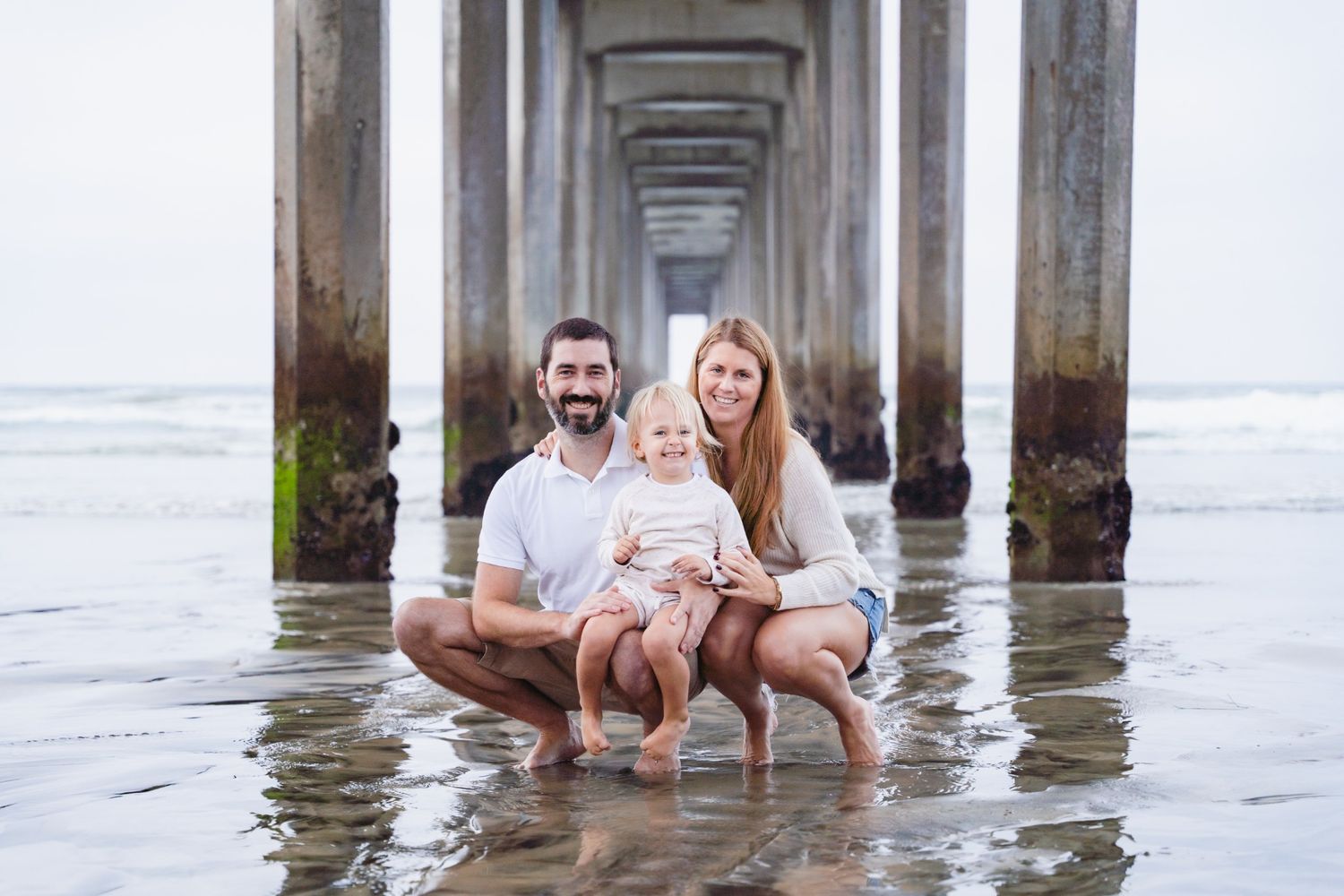 Family of three smiling under a beach pier, parents crouching with baby on a calm shoreline.