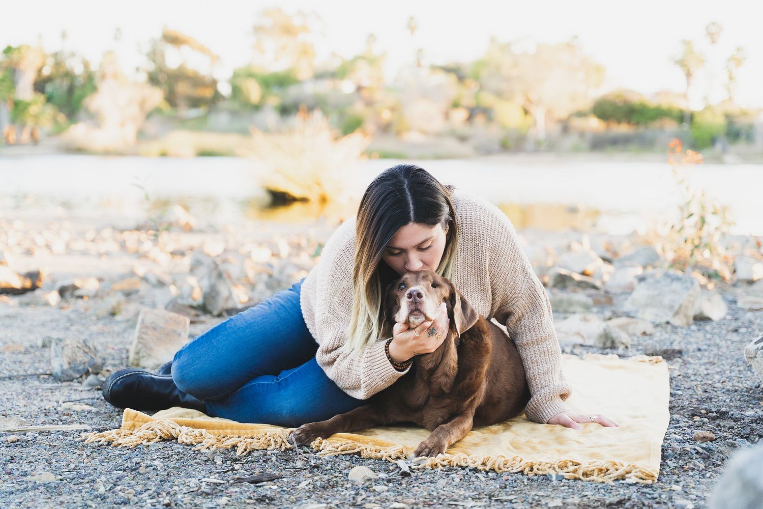 Woman in beige sweater cuddling her brown dog on a yellow blanket outdoors.
