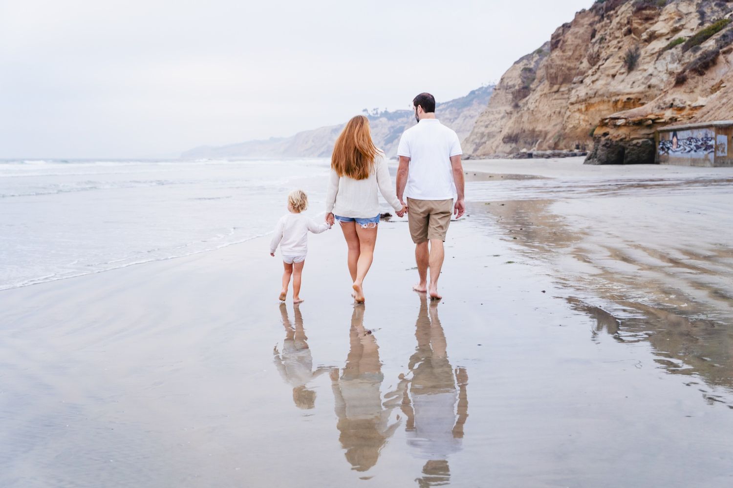 Family of three walking hand-in-hand along wet sandy beach with rocky cliffs in background on overcast day.