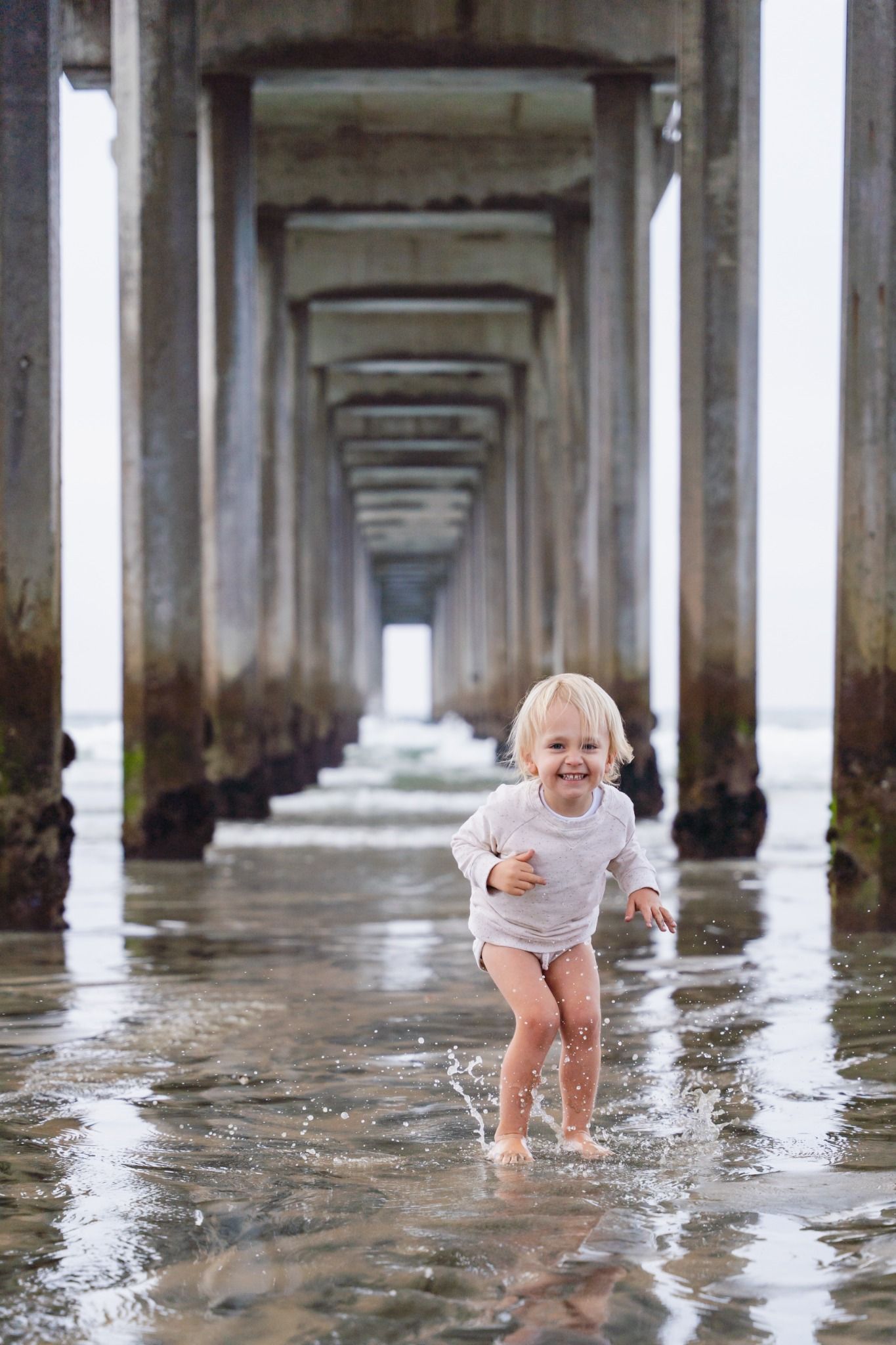 Joyful toddler splashing through shallow water beneath a wooden pier, smiling brightly at the camera.