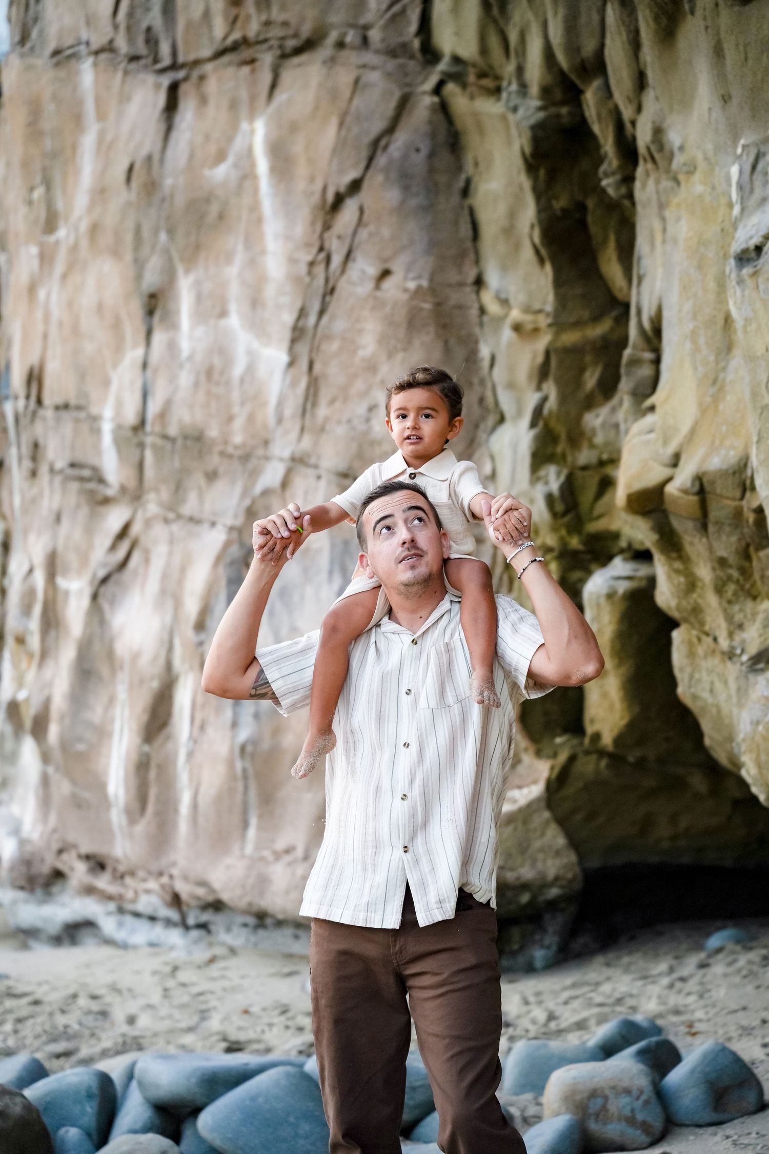 Father carrying young child on shoulders near rocky cliff at beach, smiling up at toddler in heartwarming outdoor moment.