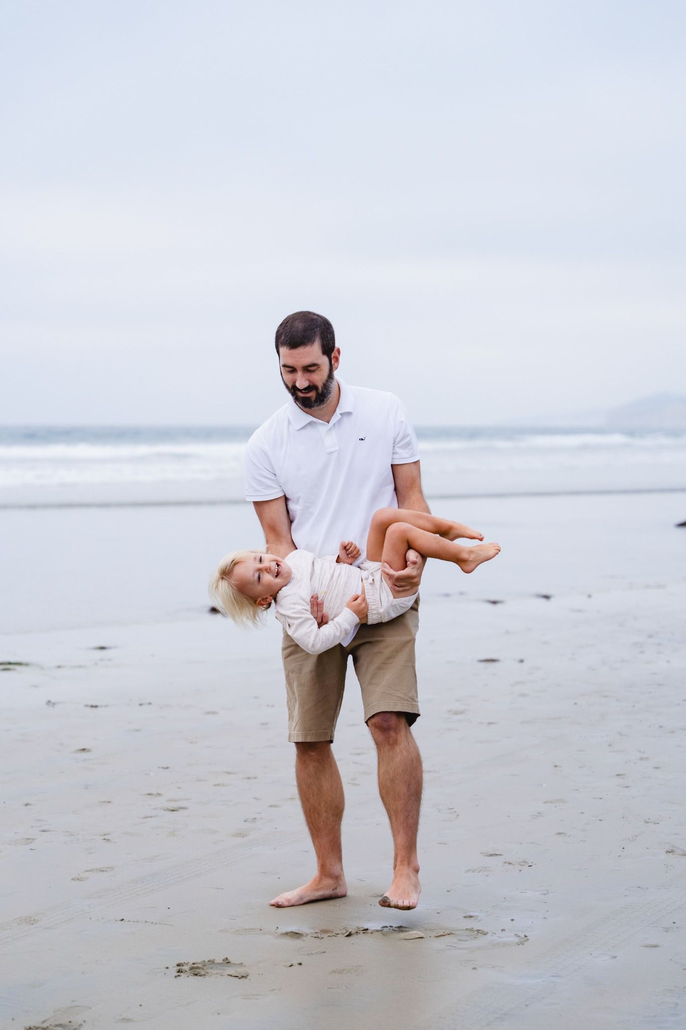 A father joyfully lifts his young blonde daughter on a sandy beach with ocean waves in the background.