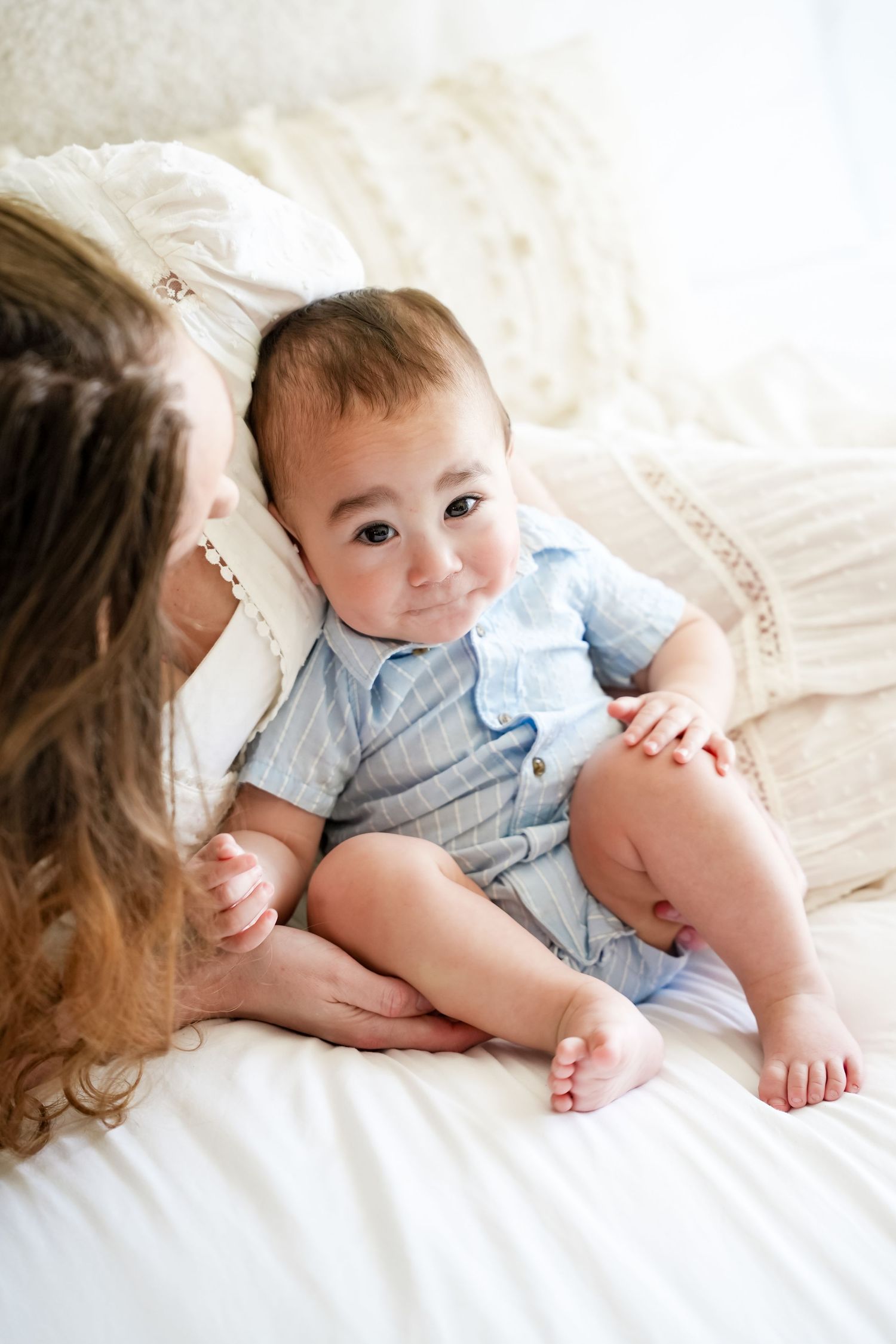 A cute baby in a blue romper sits on a white bed, looking up at a woman with curly hair.