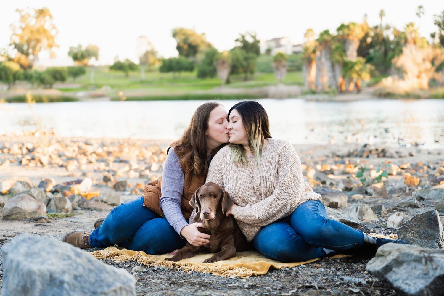 Two women kissing while sitting on rocky shore with chocolate Labrador dog on yellow blanket.