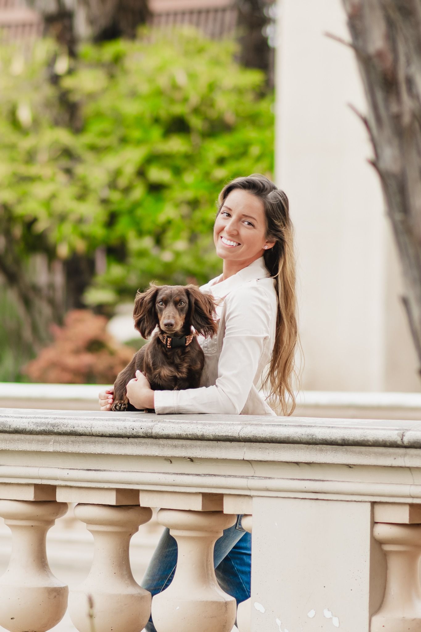 Smiling woman with long hair holding a brown dachshund on a balcony outdoors.