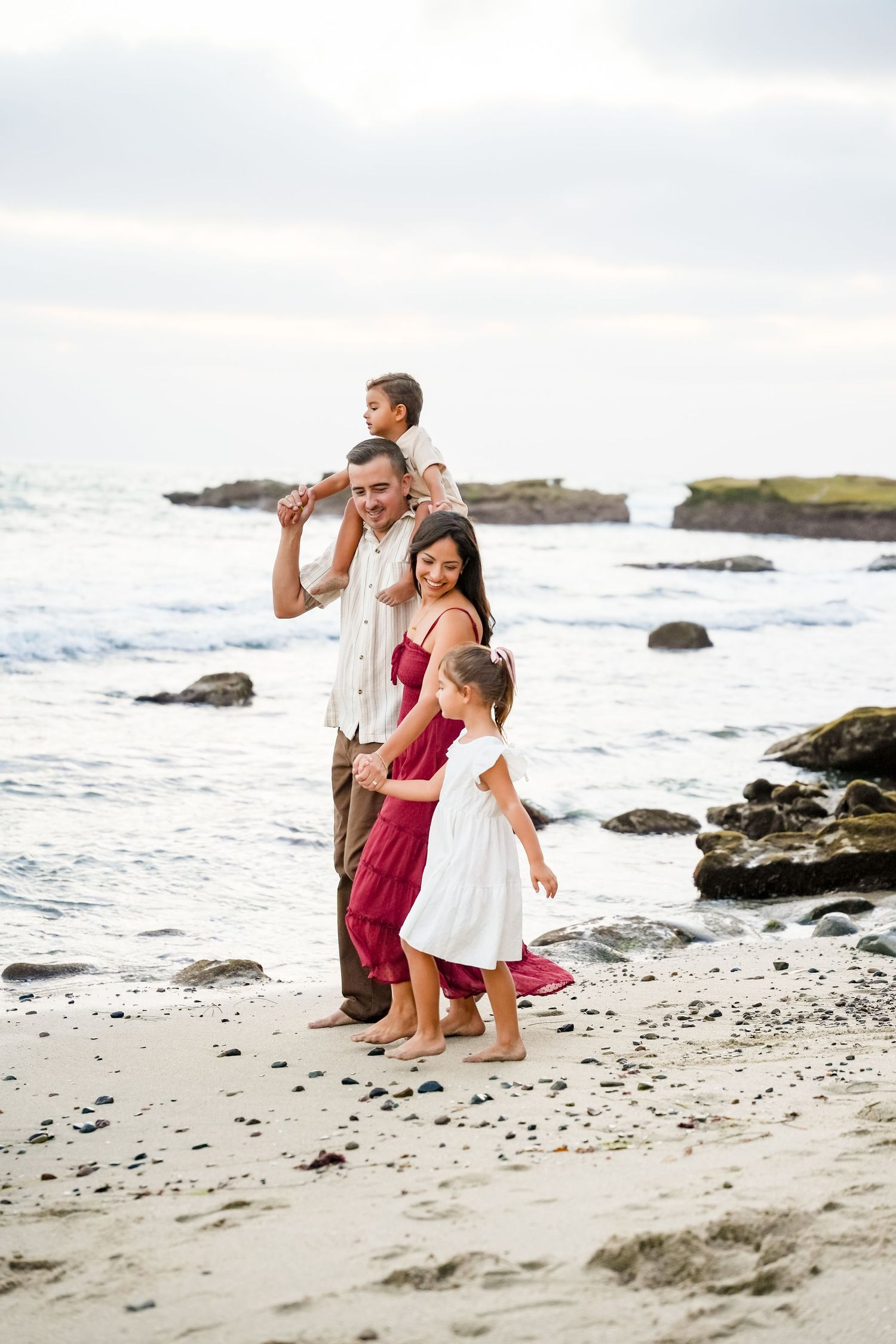 Happy family of four enjoying a barefoot walk on a rocky beach at sunset, parents and two children smiling.