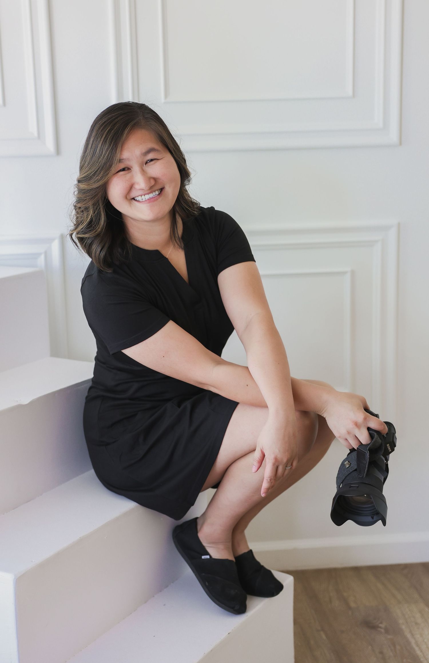 Smiling woman in black dress sitting on white stairs holding a camera.