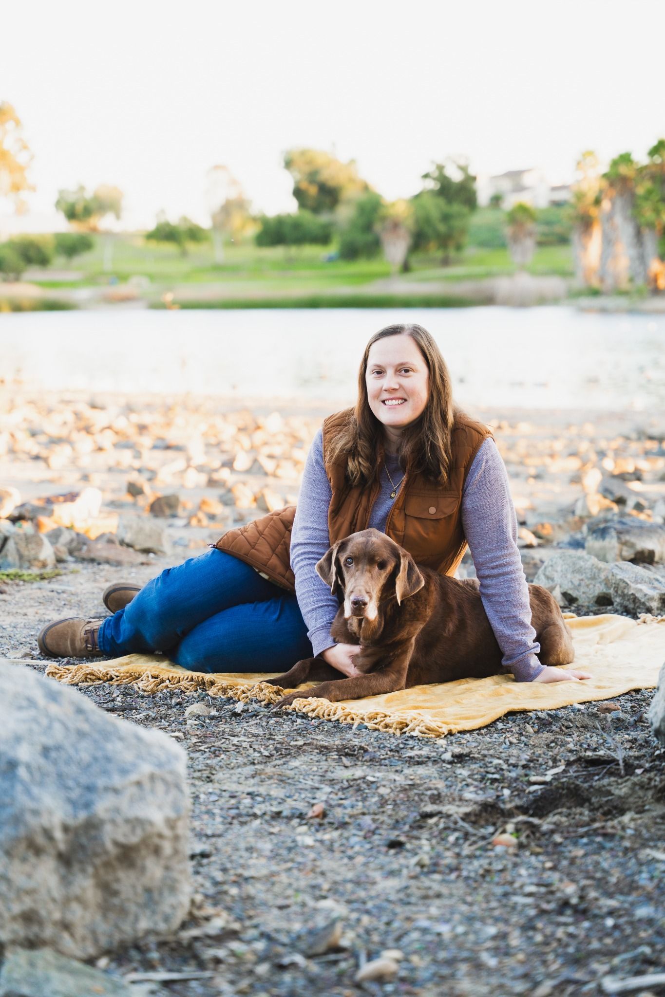 Smiling woman sitting on a yellow blanket with her chocolate Labrador Retriever on a rocky lakeside beach.