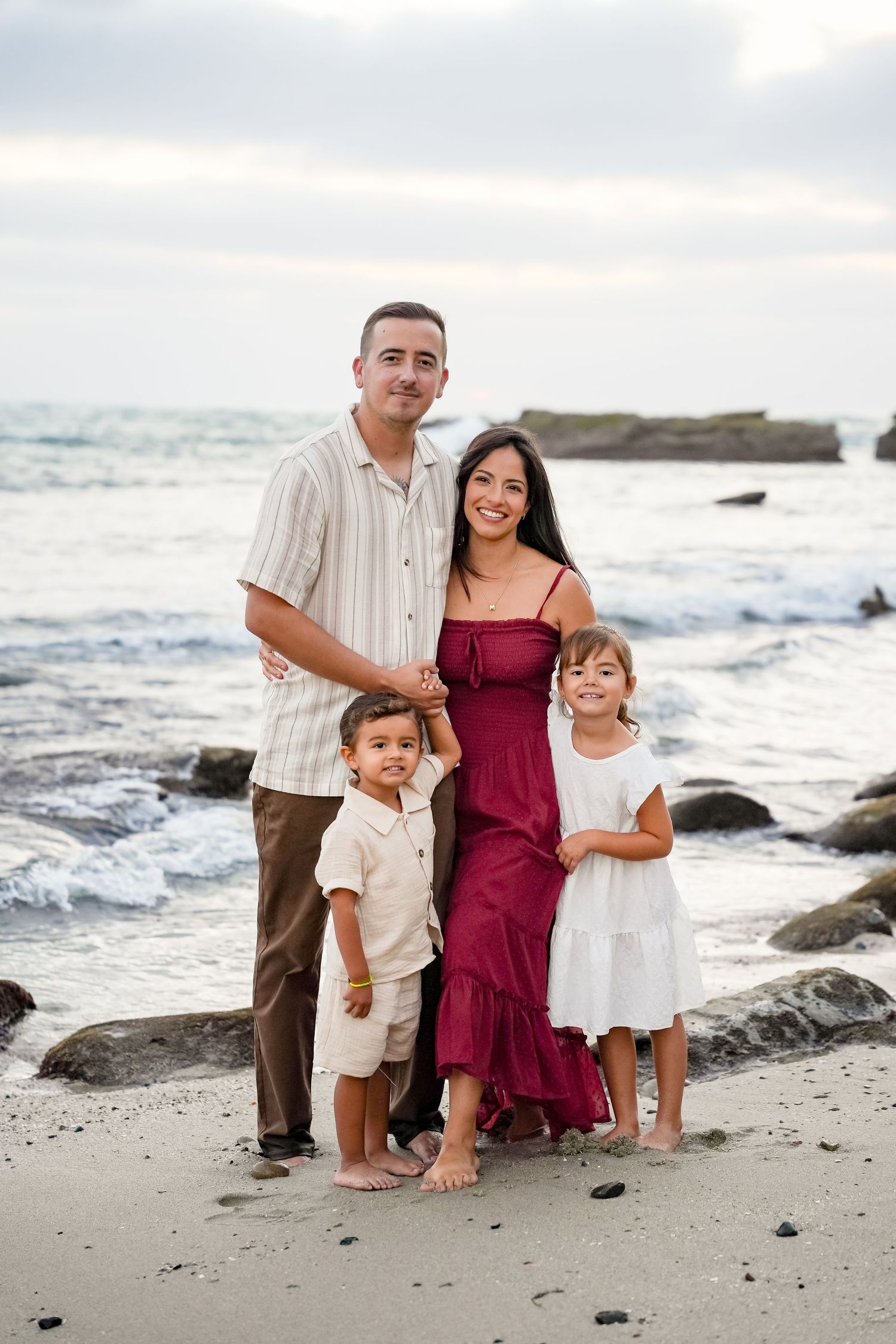 Family of four poses on a rocky beach, parents and two young children smiling, wearing coordinated outfits by the ocean.