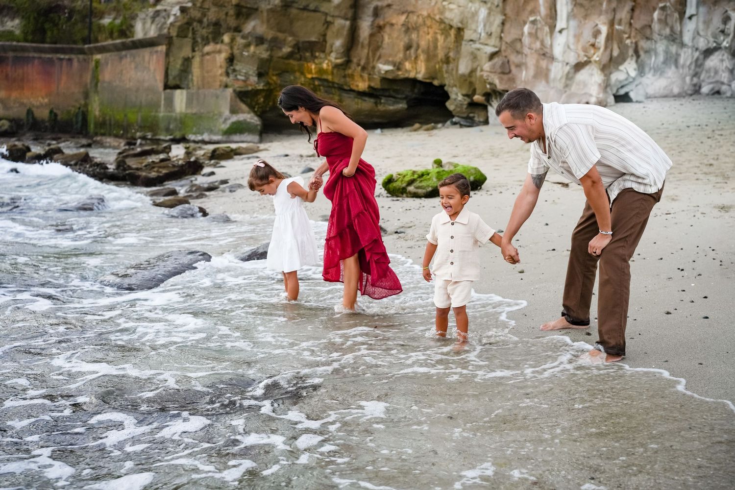 Family of four playing in ocean waves at beach, parents guiding two toddlers in white outfits, mother in red dress.