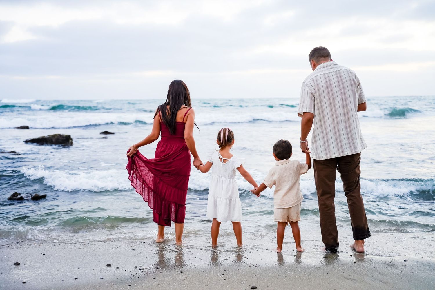 Family of four holding hands facing ocean waves on sandy beach.