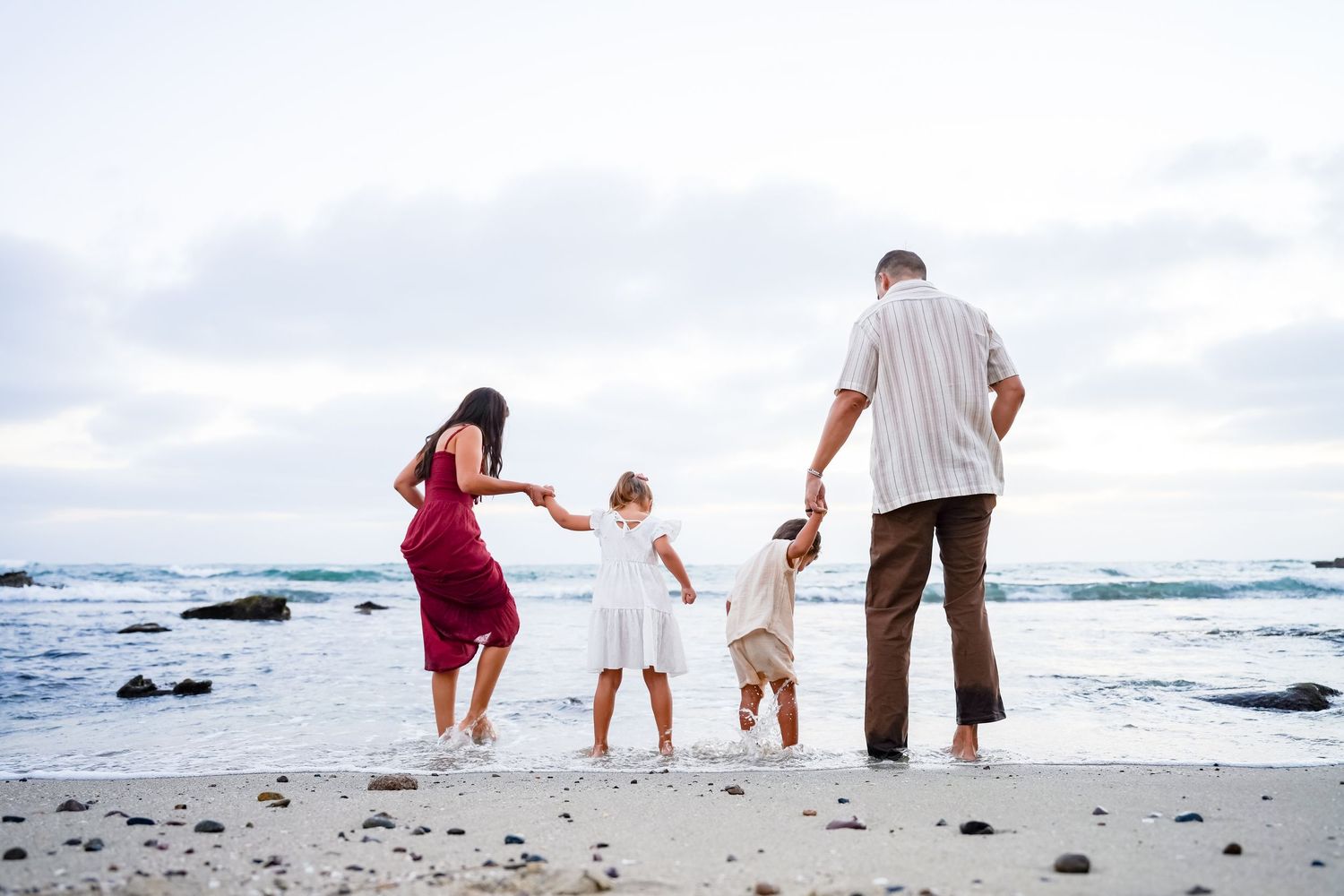 Family of four walking hand-in-hand toward ocean waves on a cloudy beach.