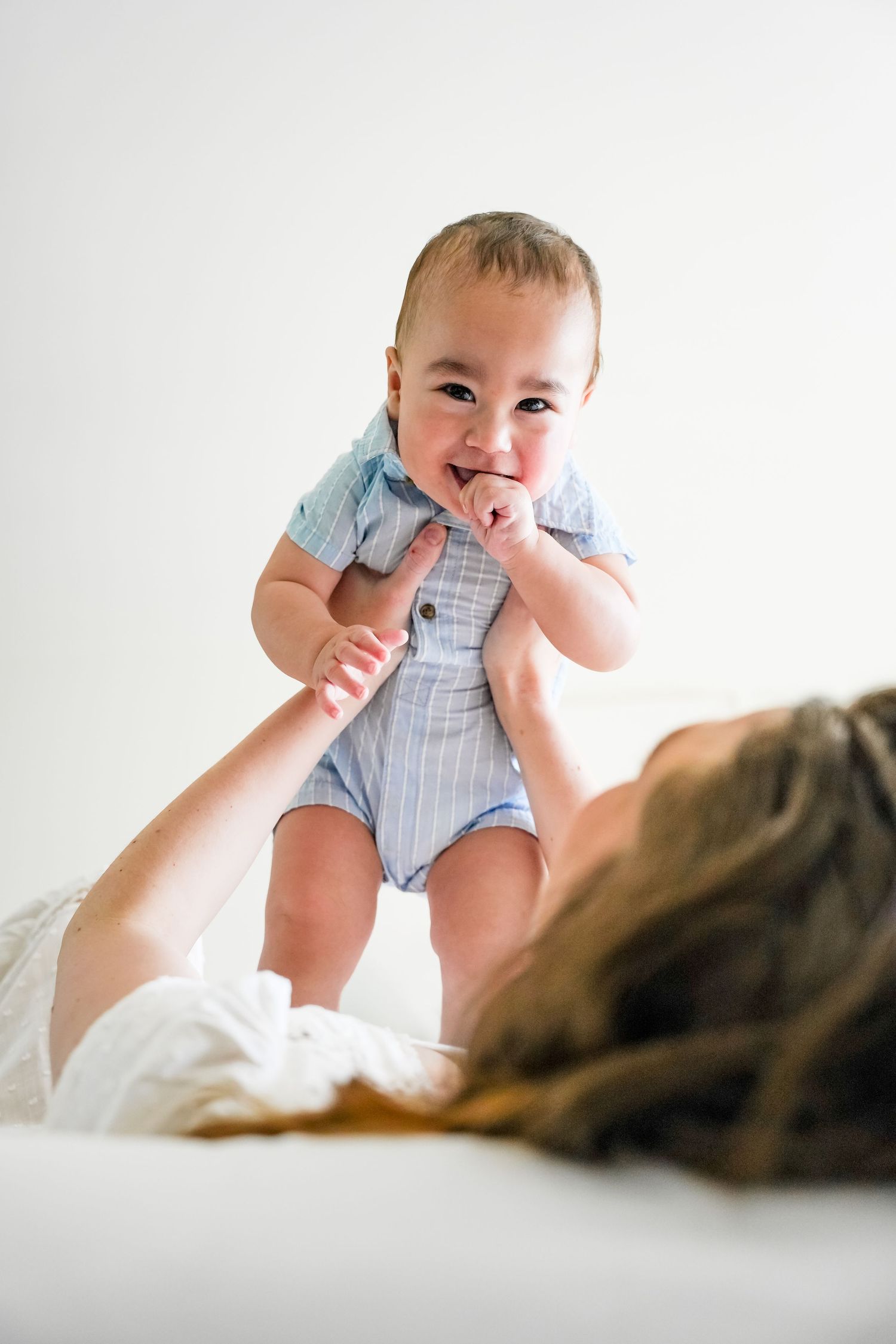 Joyful baby in blue romper being held up by mother lying on white bed, smiling and laughing.