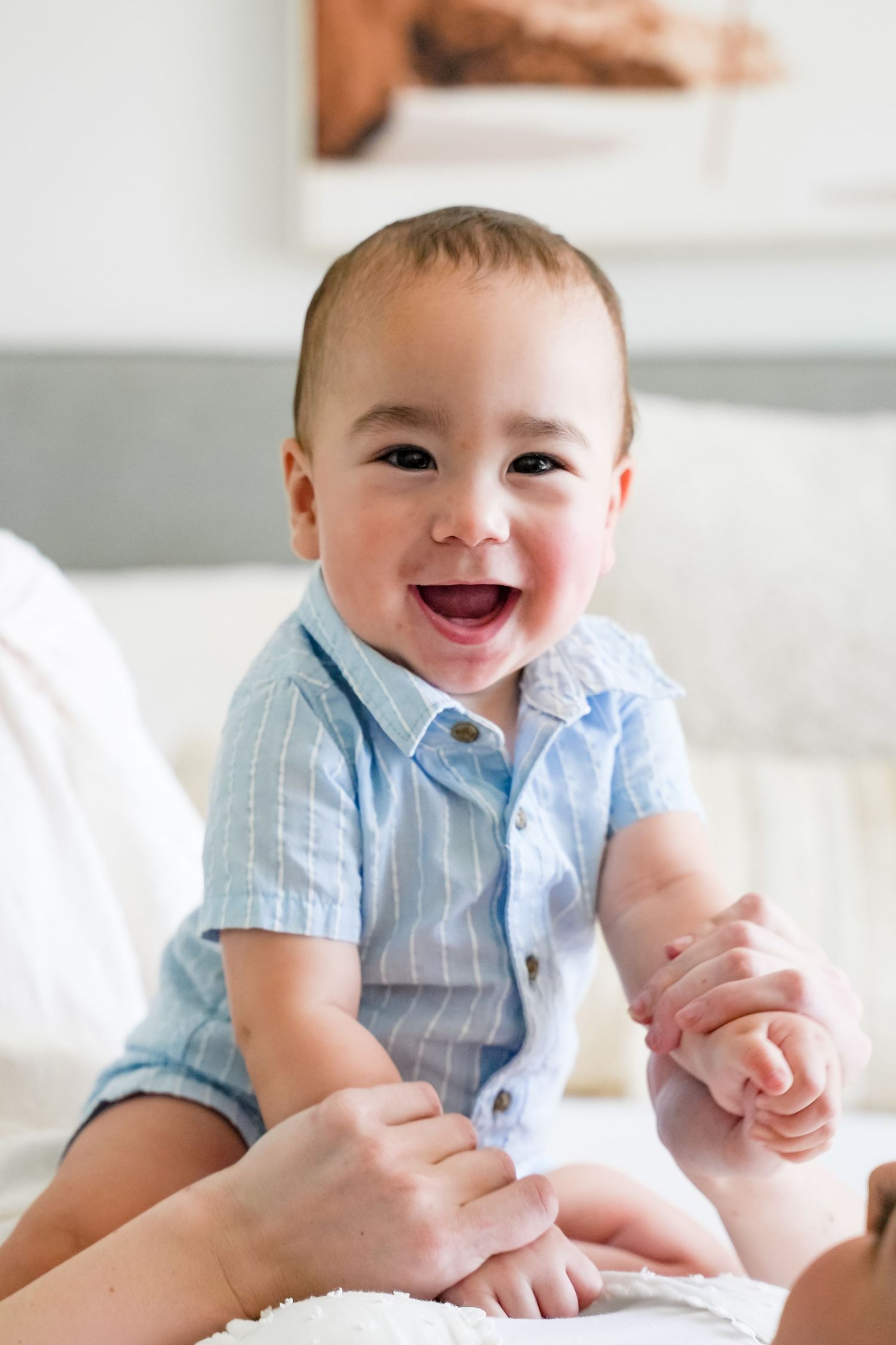 A happy baby boy in a light blue striped shirt smiles joyfully while being held by adult hands on a white bed.