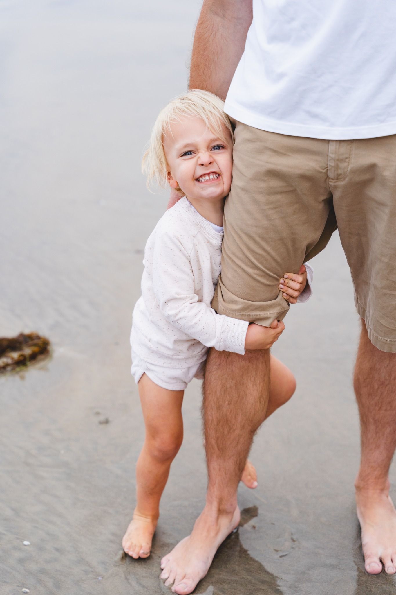 Happy blonde toddler hugging father's leg and smiling on a wet sandy beach.