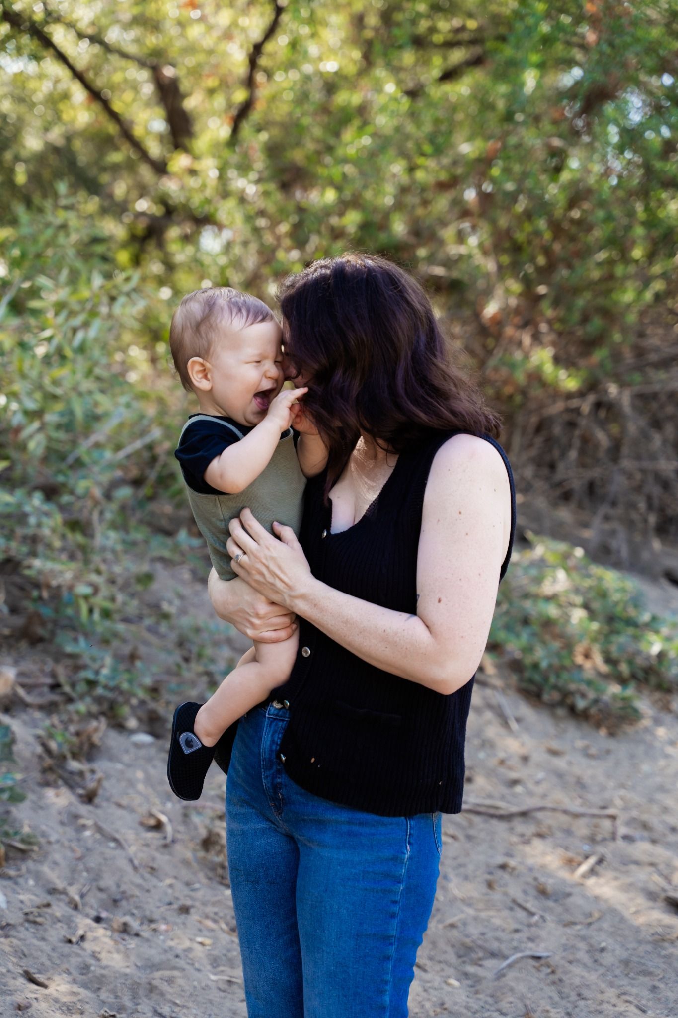 A mother holds her laughing toddler outdoors, sharing a joyful moment surrounded by lush green trees.